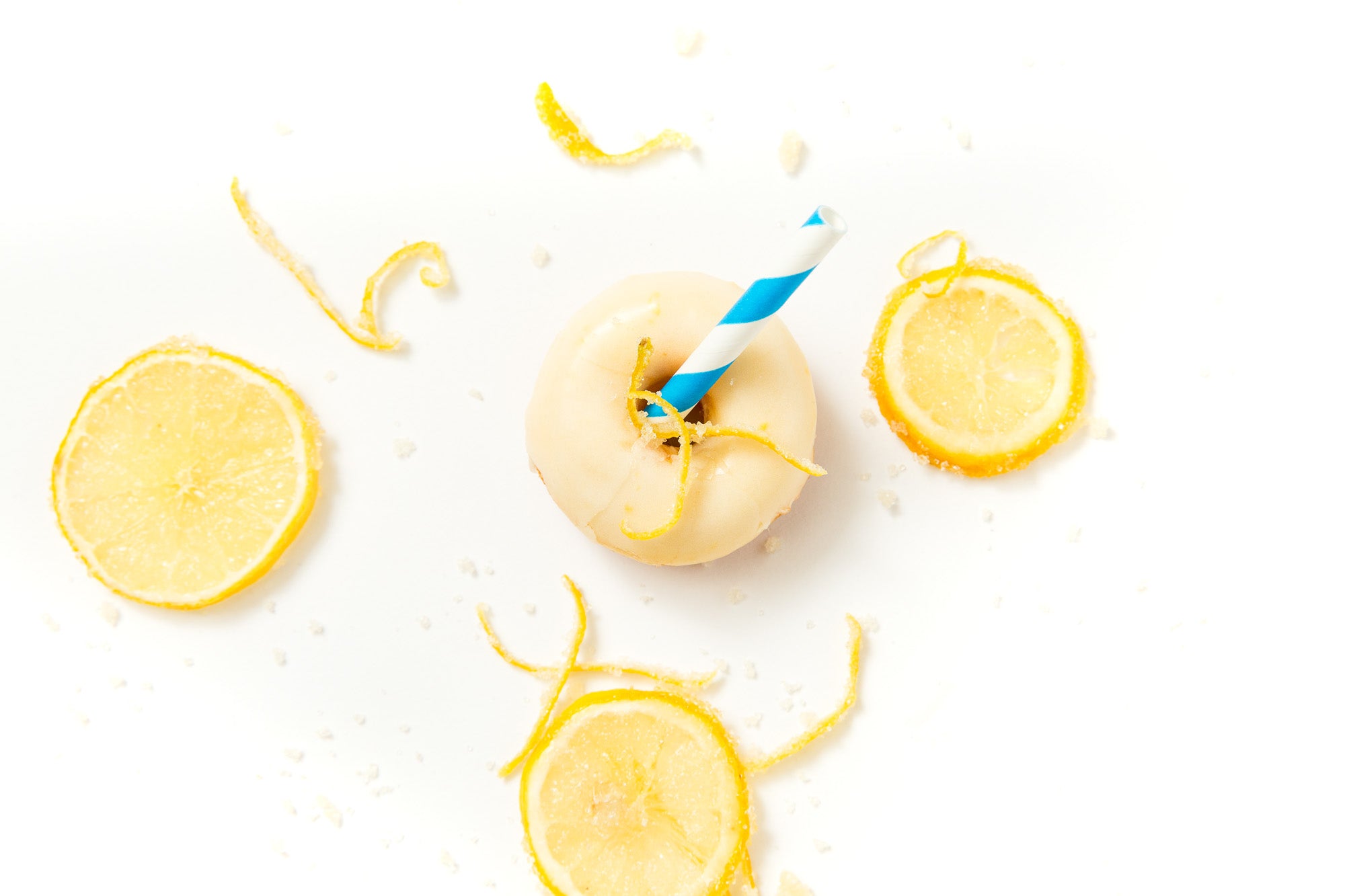 Image from above of a Miss Jones Baking Co Lemonade Donut with a blue and white straw in the center, surrounded by candied lemon slices, zested lemon and sugar