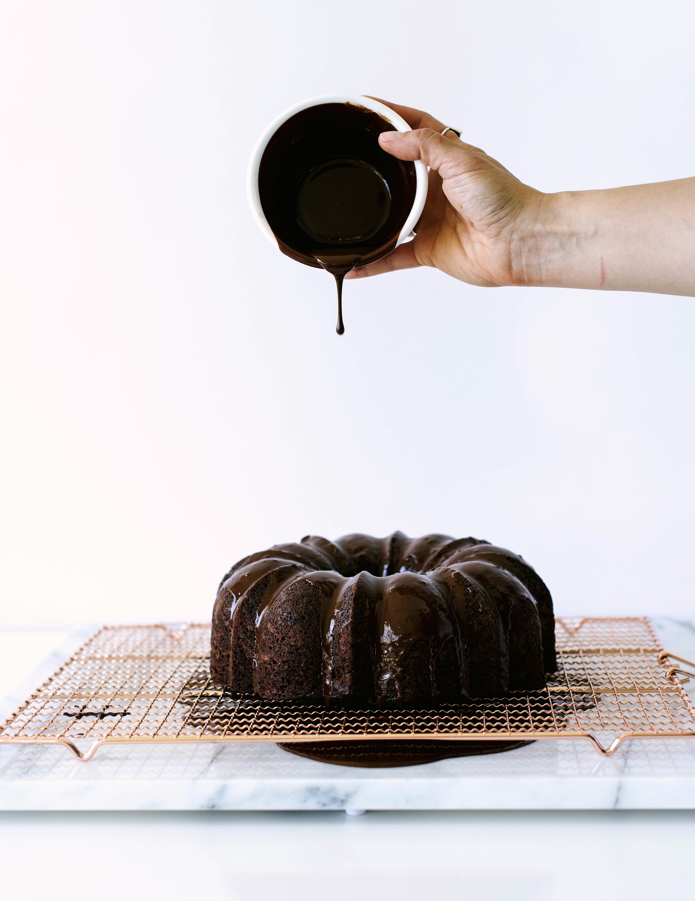 A hand pouring chocolate ganache on the top of Miss Jones Dark Chocolate Coconut Bundt Cake on a baking sheet