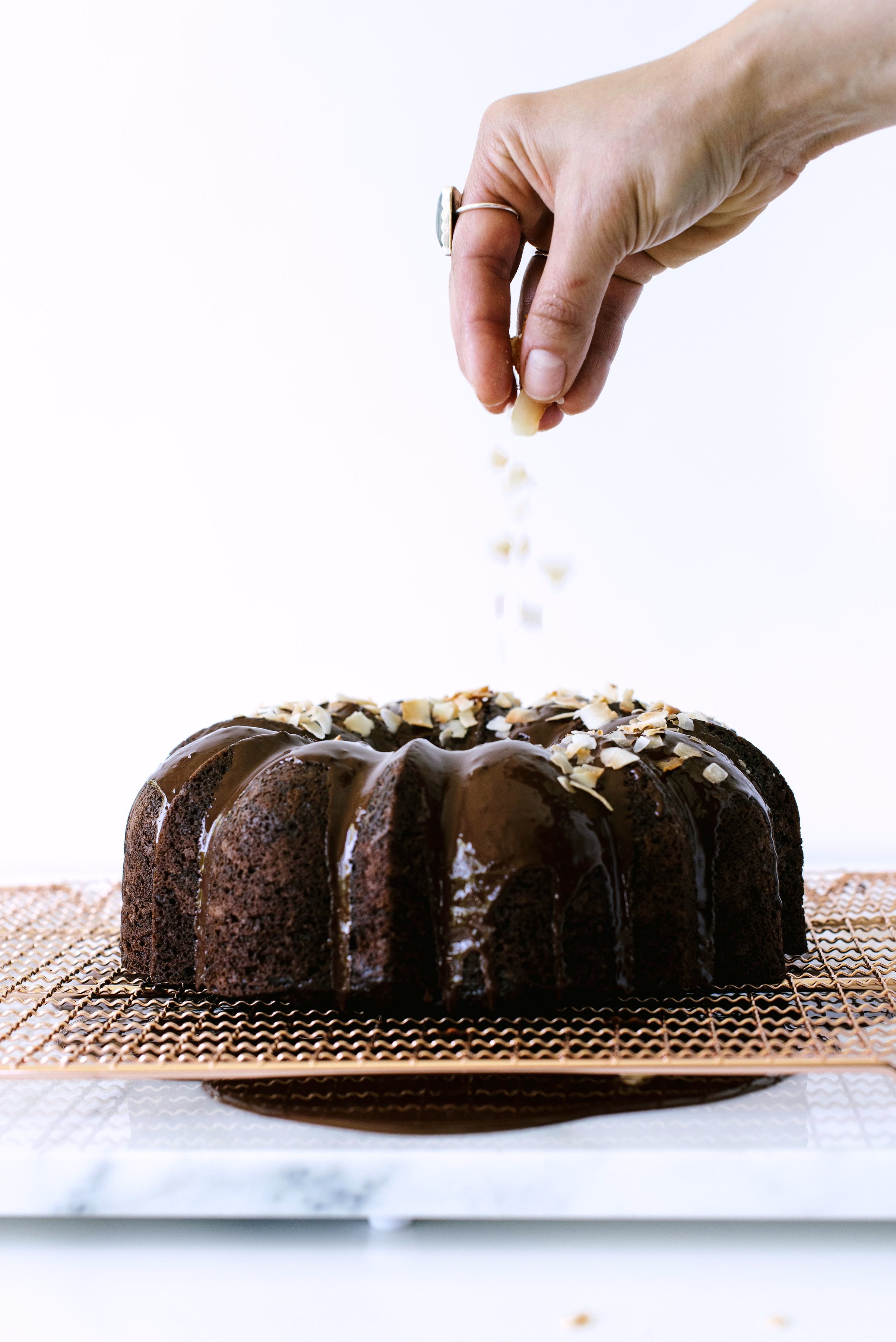 A hand sprinkling toasted coconut flakes on the top of Miss Jones Dark Chocolate Coconut Bundt Cake on a baking sheet