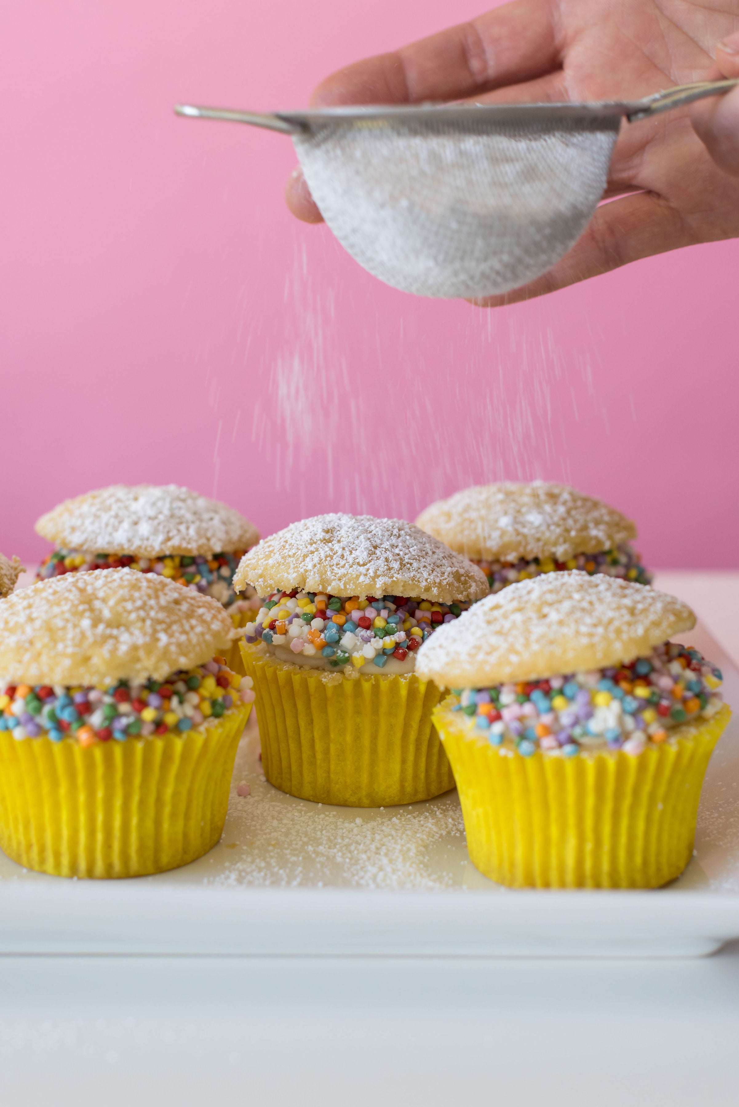 Image of side of five Miss Jones Baking Co Confetti Pop Cream Puff Cupcakes being sprinkled with powdered sugar