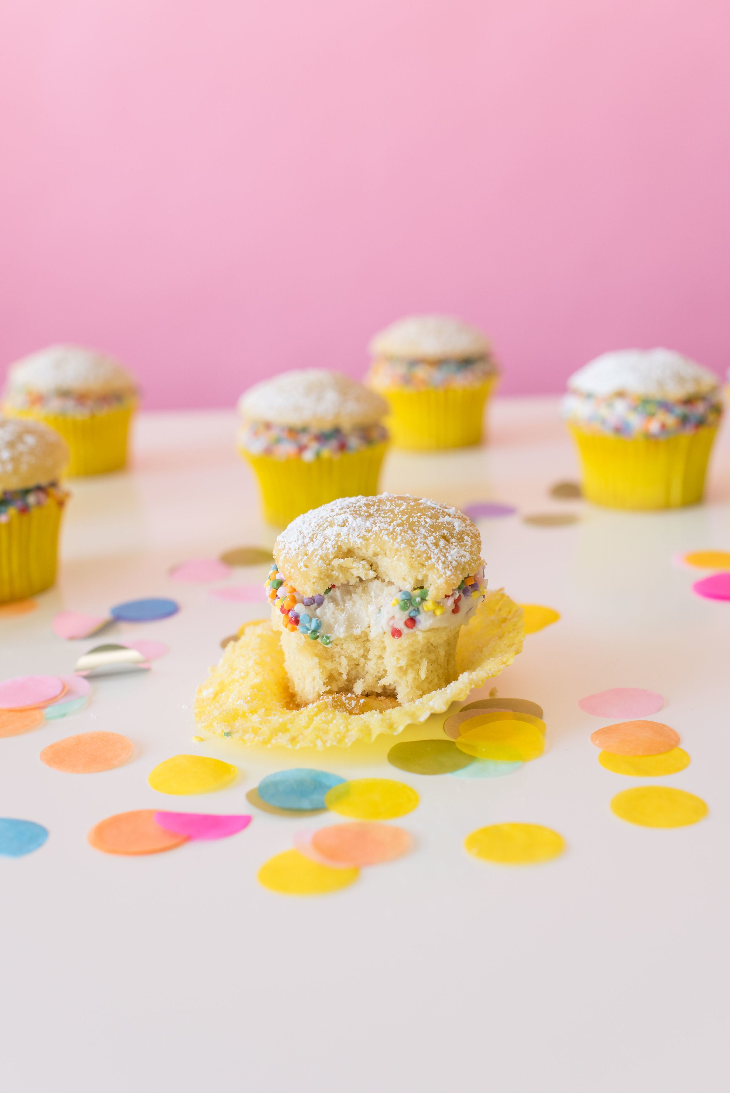 Image of side of six Miss Jones Baking Co Confetti Pop Cream Puff Cupcakes with a bite taken out of middle cupcake