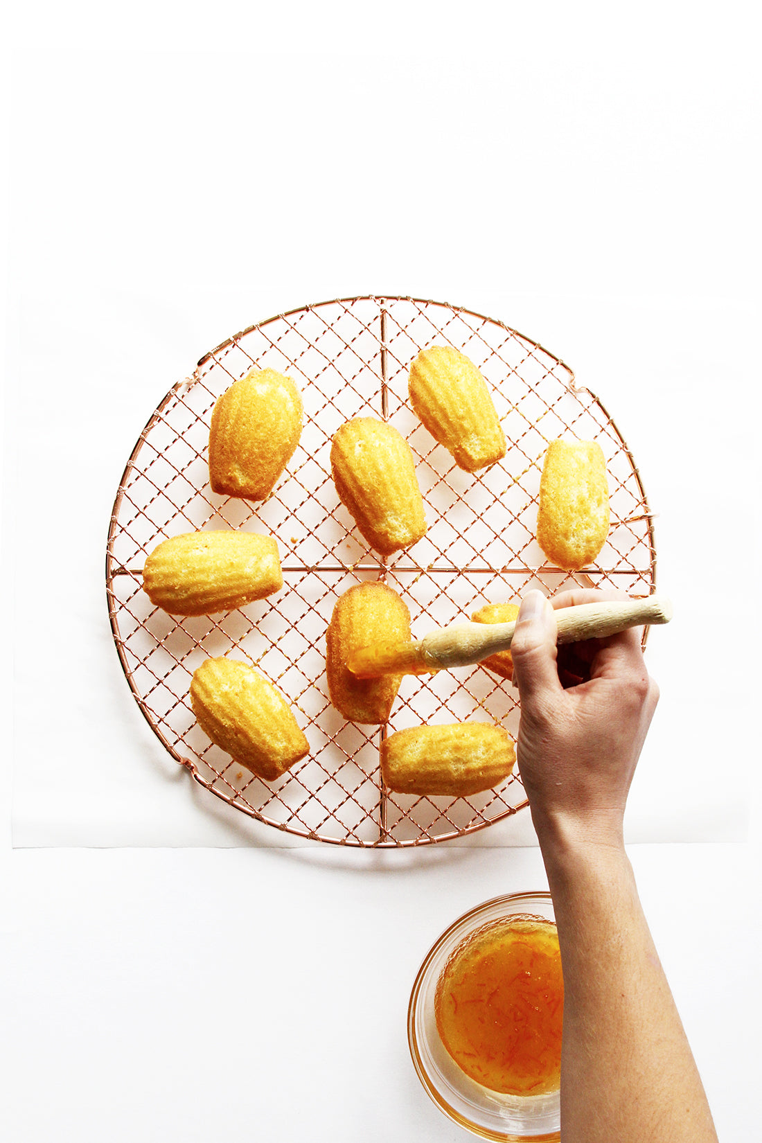 Image of nine Miss Jones Baking Co Marmalade Madeleines on a baking rack being glazed with orange marmalade by a hand