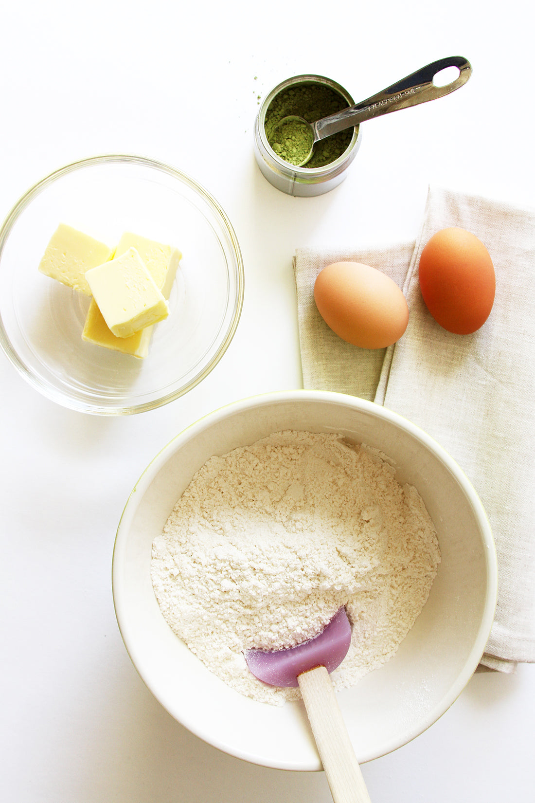 Image from above of a measuring spoon in a jar of powdered matcha next to two eggs, a bowl of butter slices, and a bowl of cake mix for Miss Jones Baking Co Matcha Tea Cakes