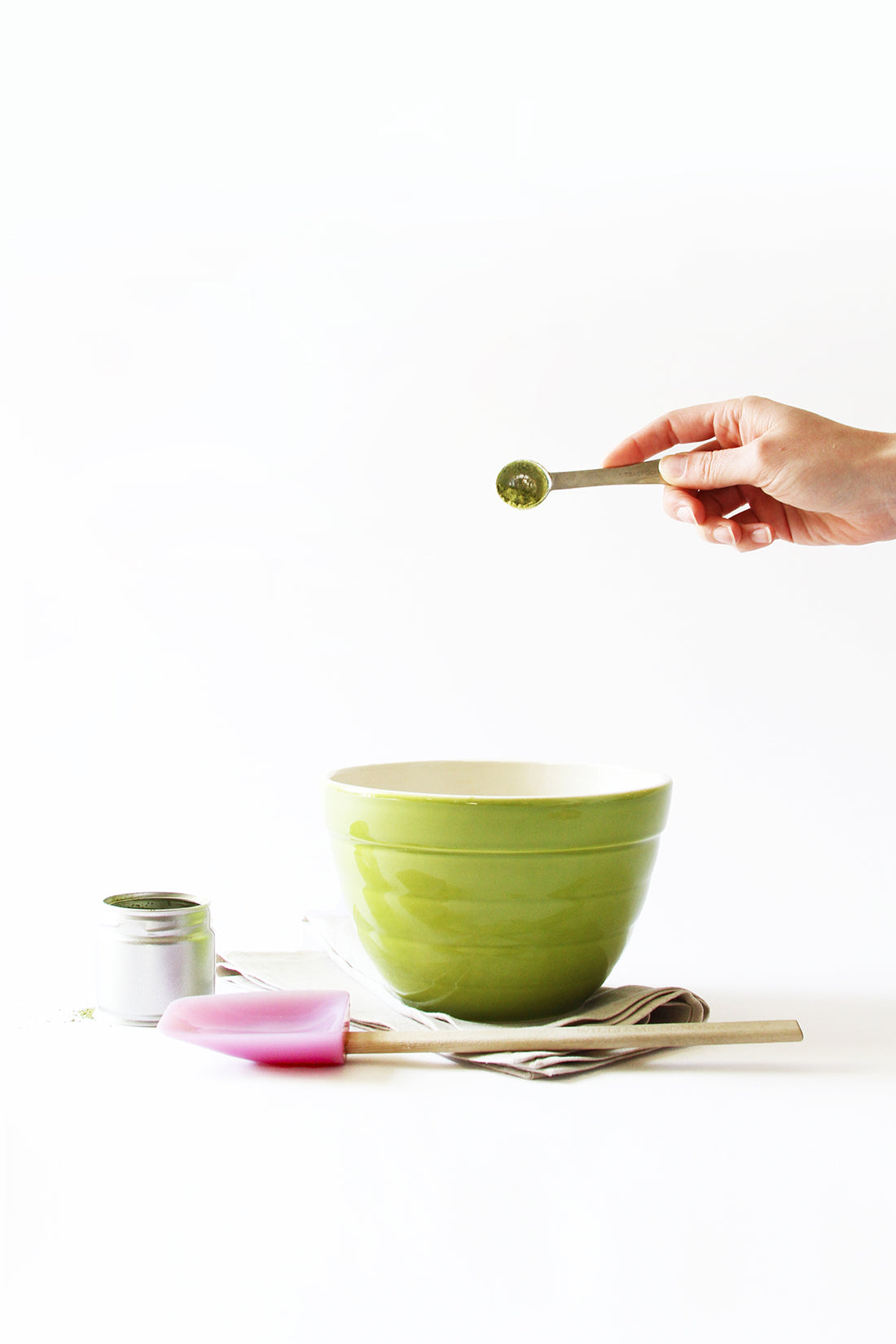 Image from the side of a spoon of matcha poured into a green mixing bowl next to a jar of powdered matcha and a purple spatula for Miss Jones Baking Co Matcha Tea Cakes