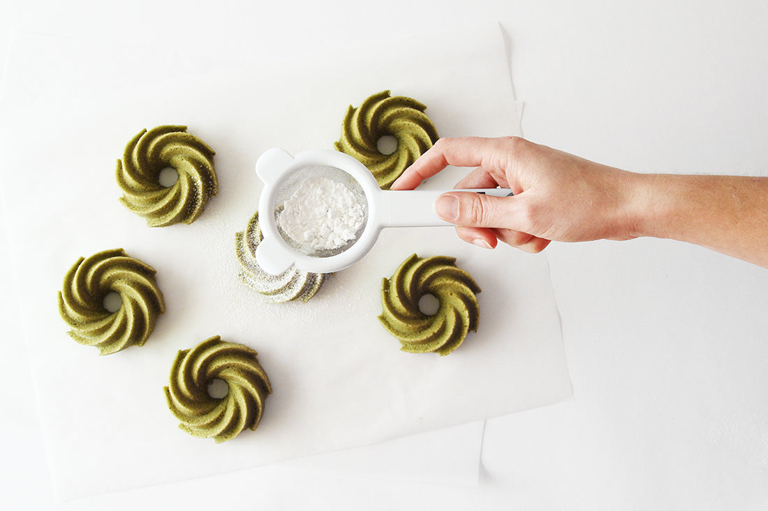 Image from above of a hand putting powdered sugar on top of six Miss Jones Baking Co Matcha Tea Cakes