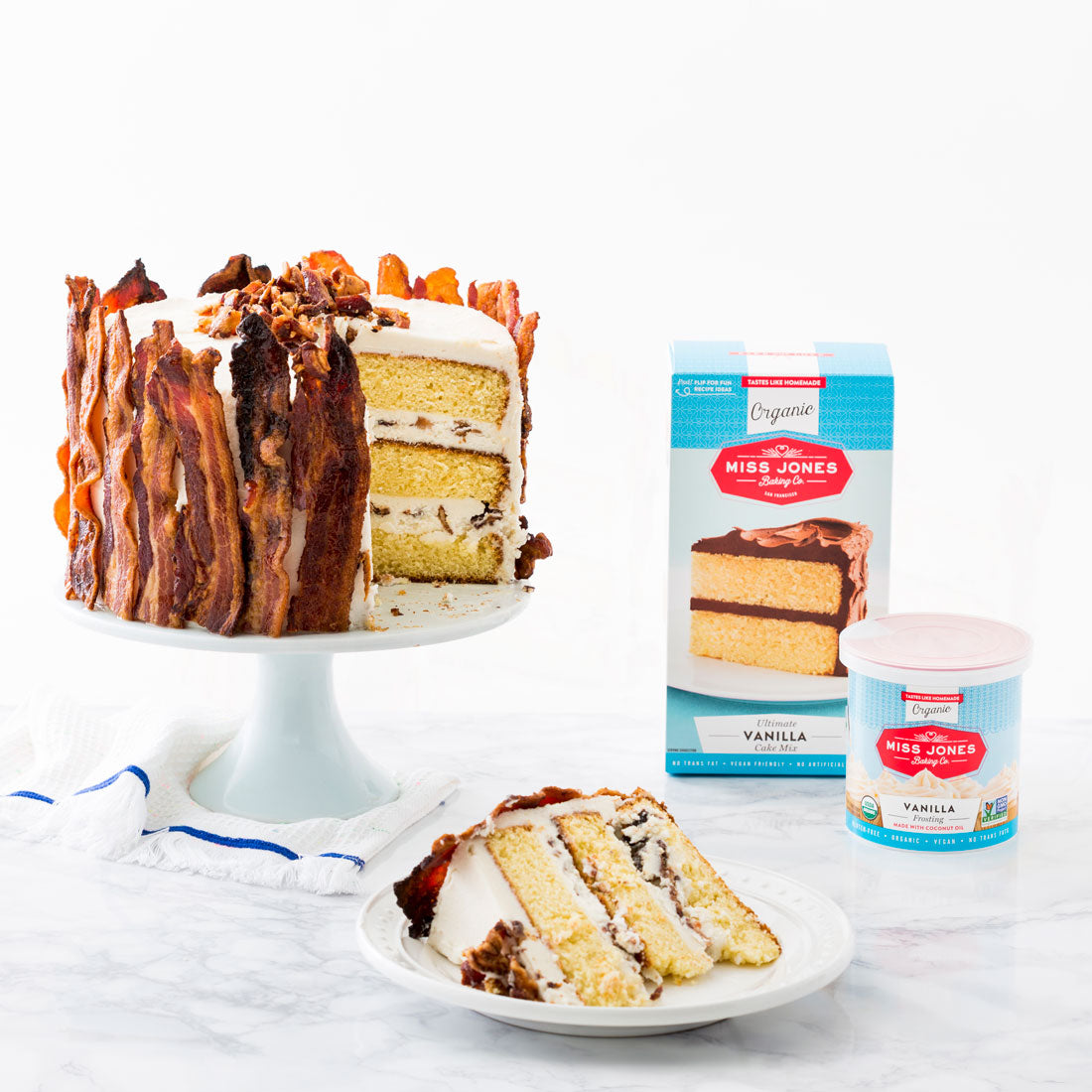 Image of side of Miss Jones Baking Co Epic Father's Day 3-Layer Bacon and Beer Cake on a cake stand with a slice on a plate below, next to a box of Miss Jones Vanilla Cake Mix and a jar of Miss Jones Vanilla Frosting