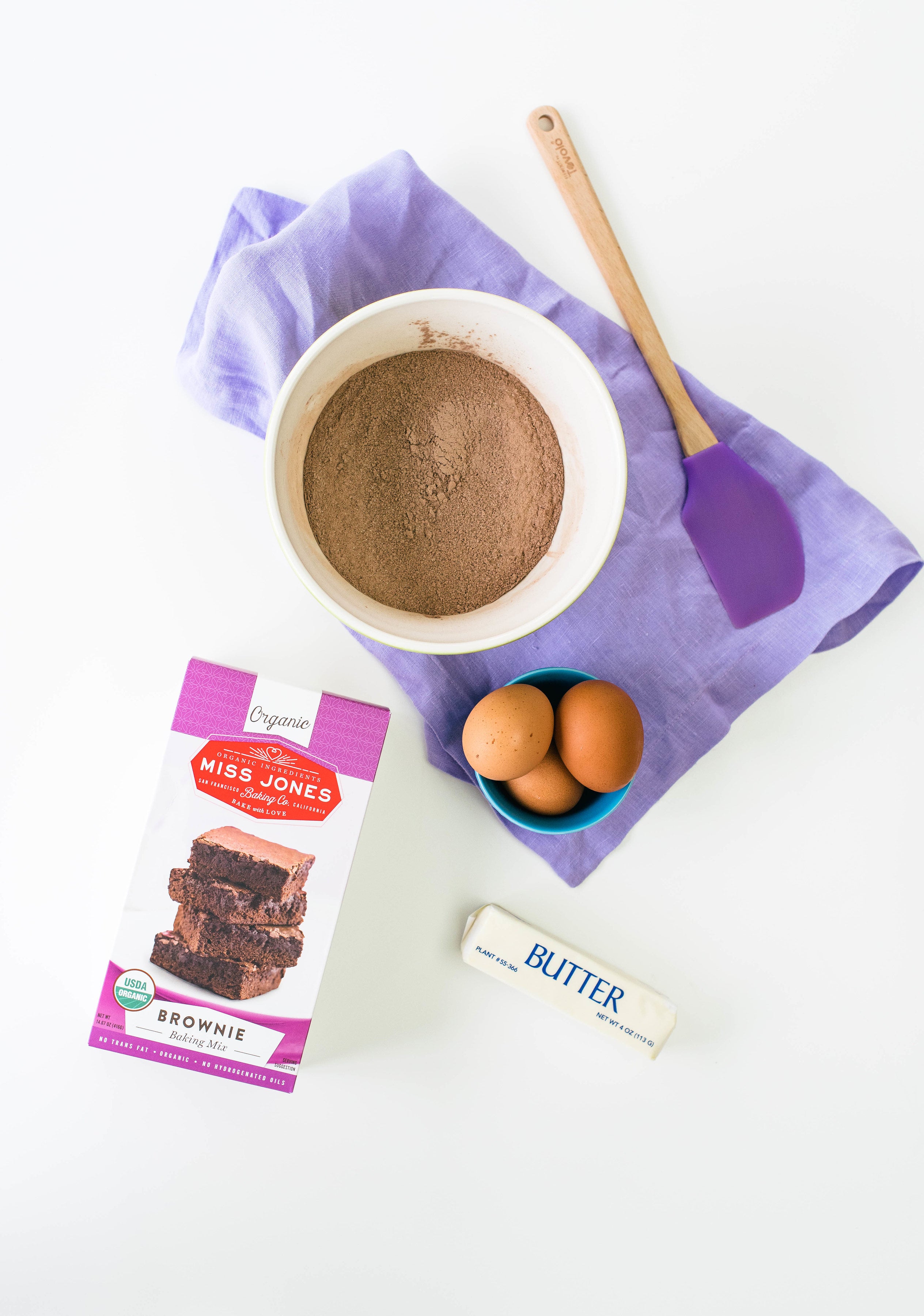 Image from above of a mixing bowl with Miss Jones Brownie Mix next to a box of Miss Jones Brownie Mix, a stick of butter, a bowl with three eggs and a purple spatula for Miss Jones Baking Co Salted Caramel Brownie Pie recipe