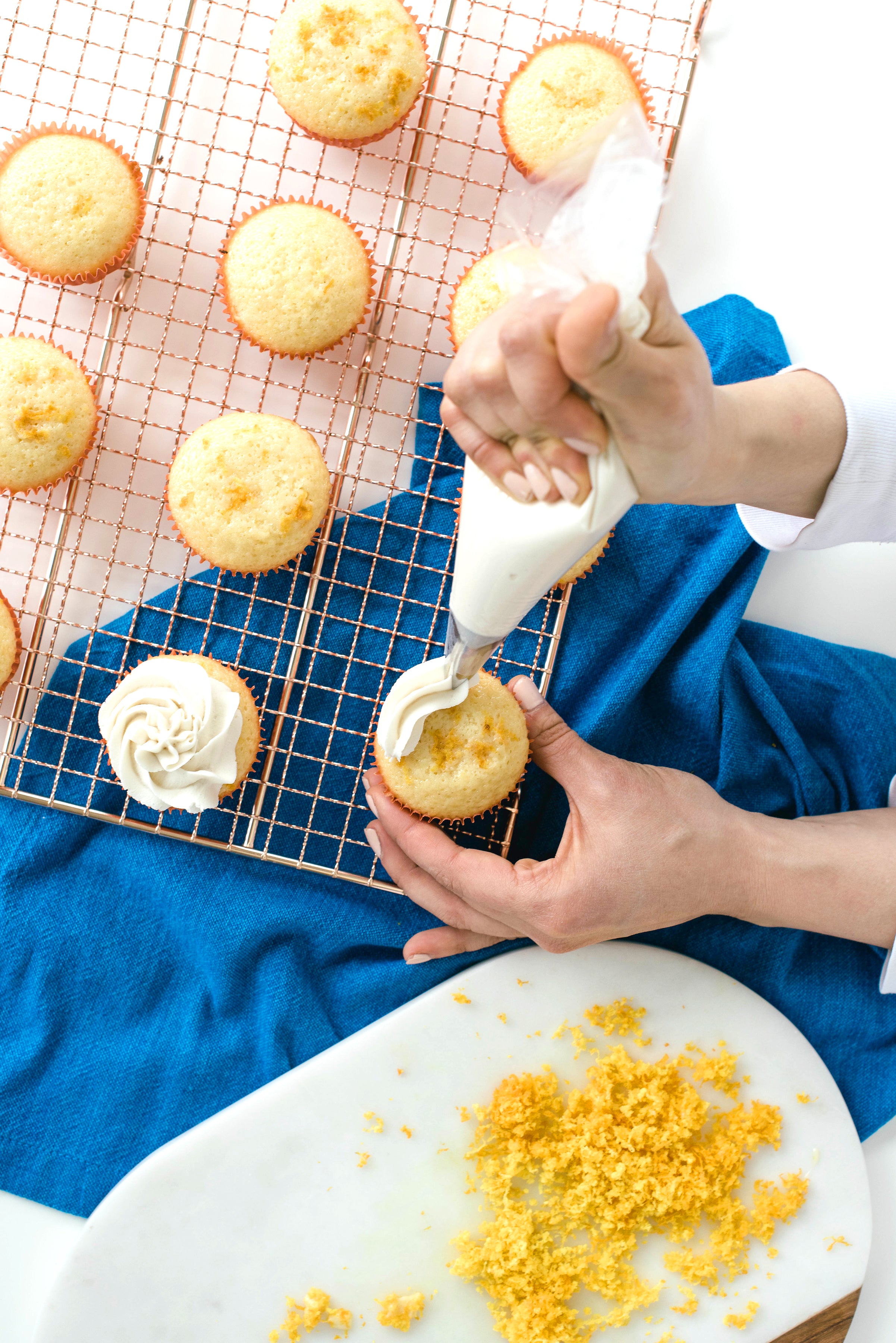 Image from above of Miss Jones Baking Co Mimosa Cupcakes on a baking rack being frosted by a hand next to orange zest on a cutting board