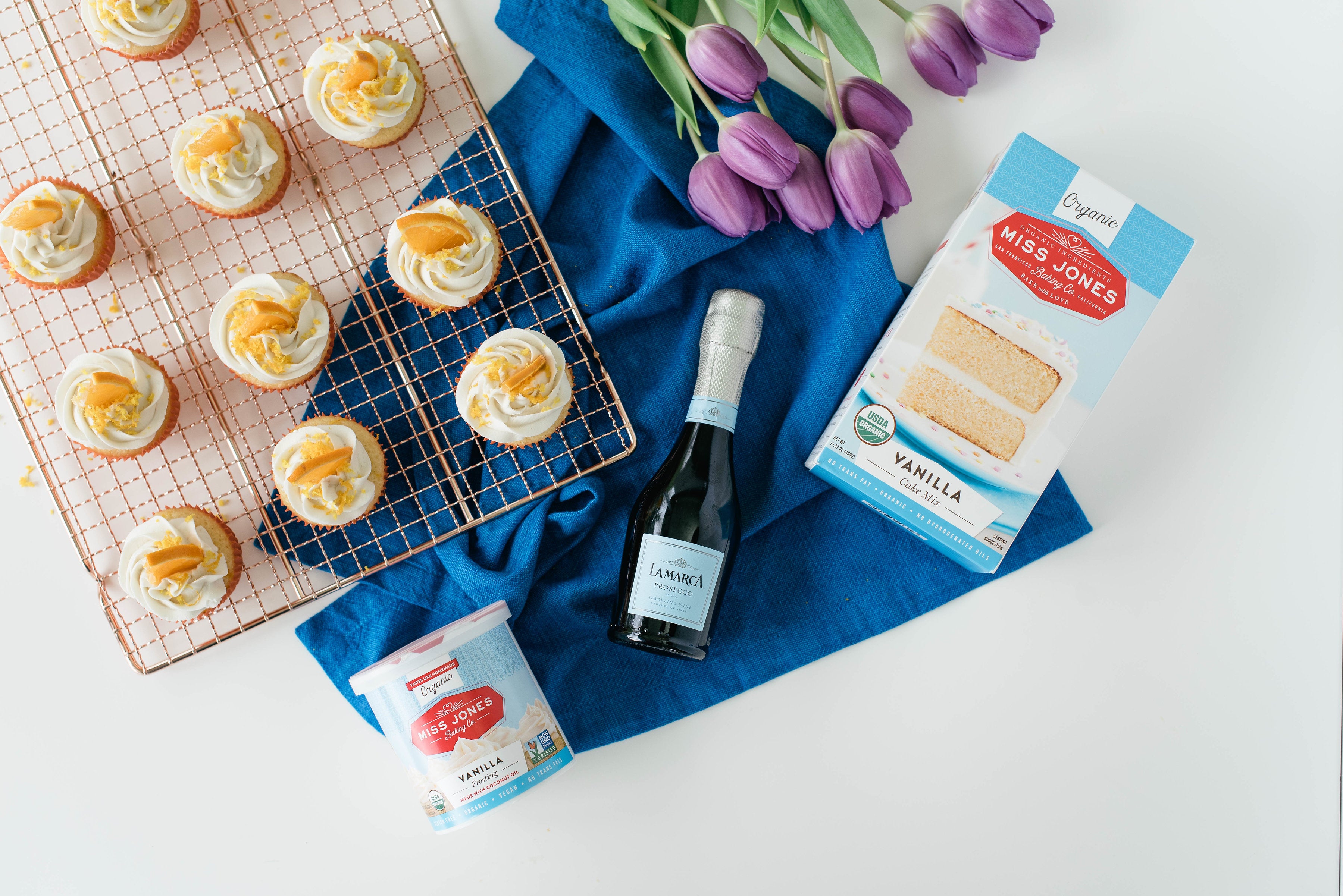 Image from above of Miss Jones Baking Co Mimosa Cupcakes on a baking rack, next to a jar of Miss Jones Vanilla Frosting, a box of Miss Jones Vanilla Cake Mix, a bottle of La Marca champagne and purple tulips