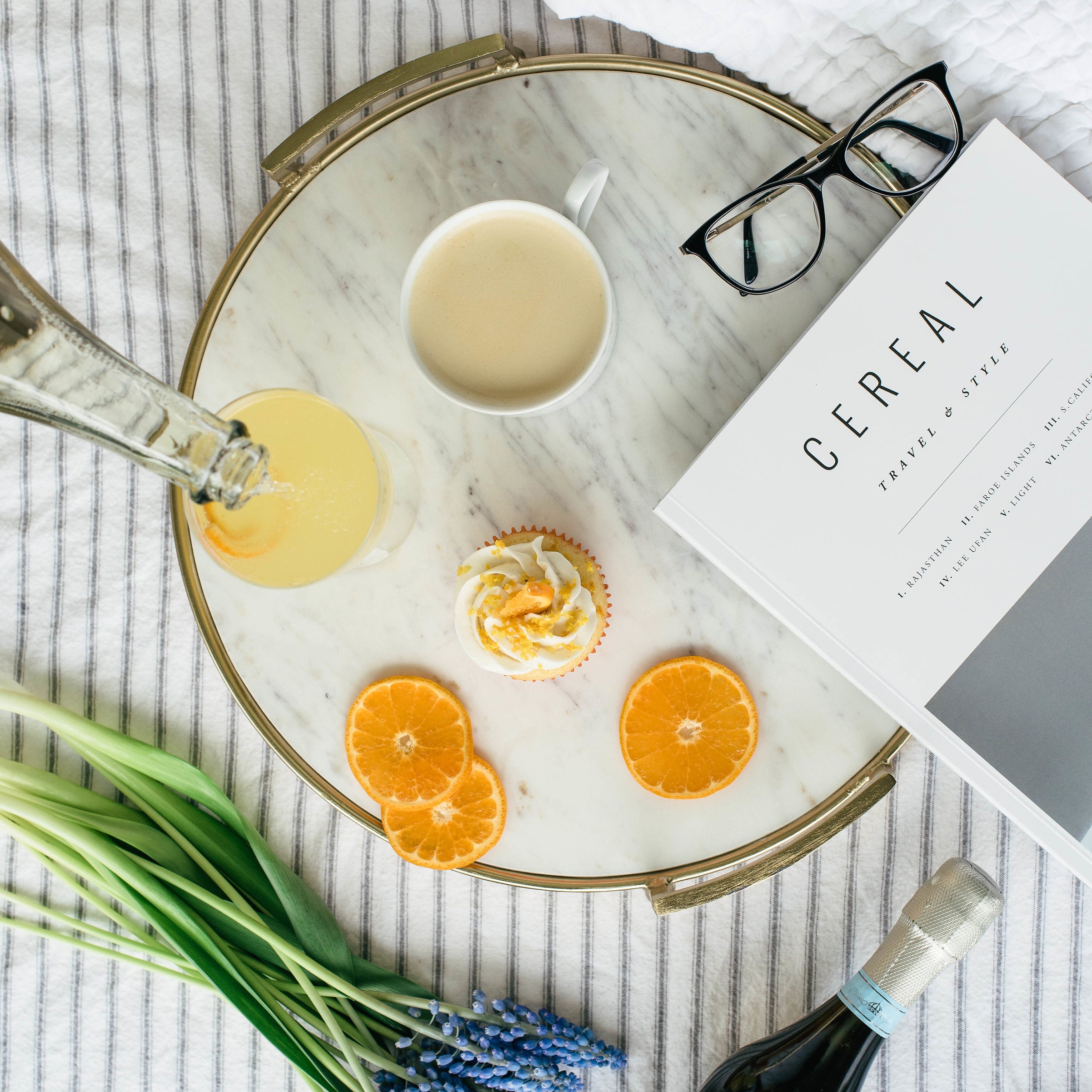 Image from above of a Miss Jones Baking Co Mimosa Cupcake on a platter next to orange slices, a mimosa, a cup of tea, reading glasses, a magazine, lavender and a bottle of champagne