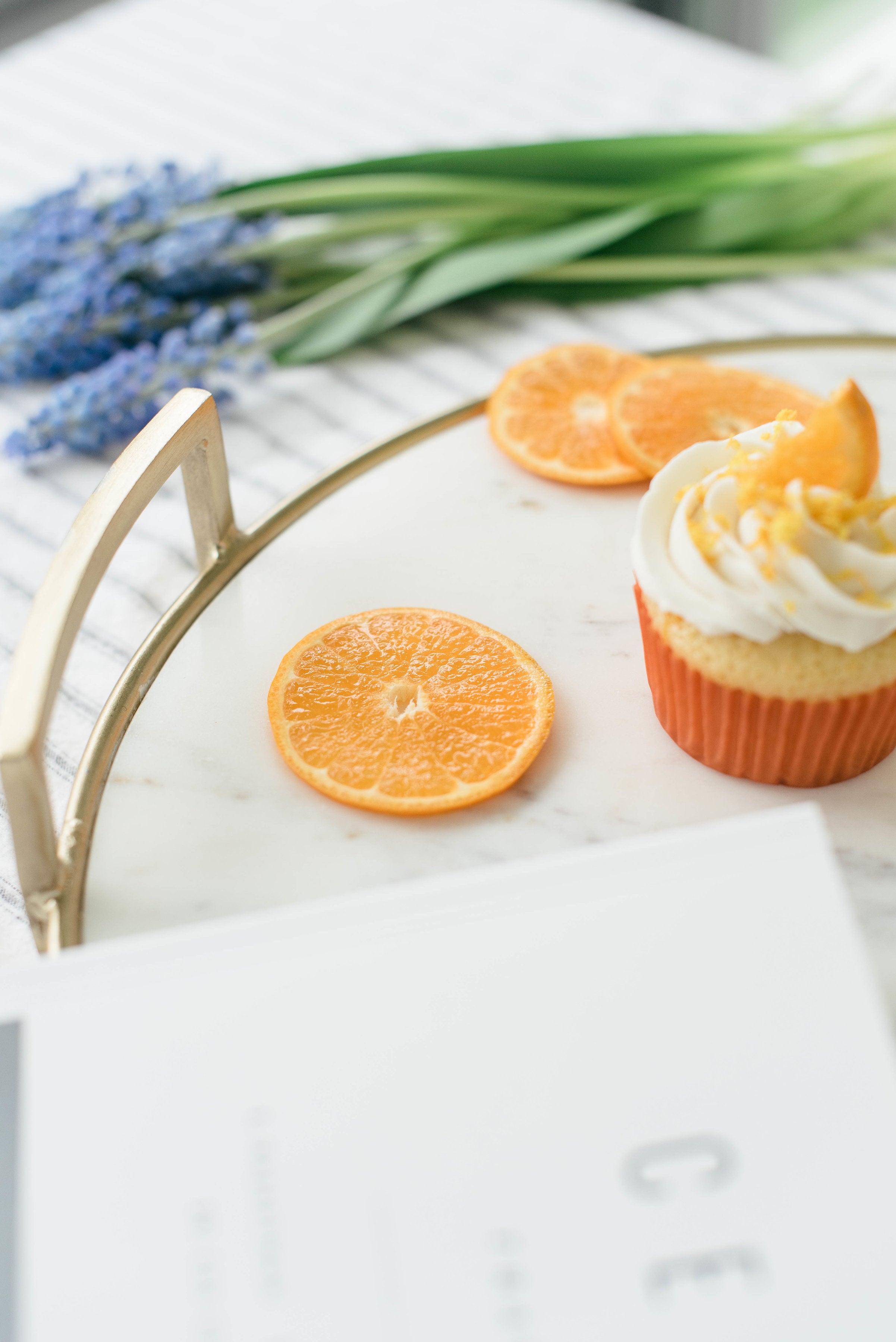 Close up image of the side of a Miss Jones Baking Co Mimosa Cupcake on a platter next to orange slices, a magazine, and a bouquet