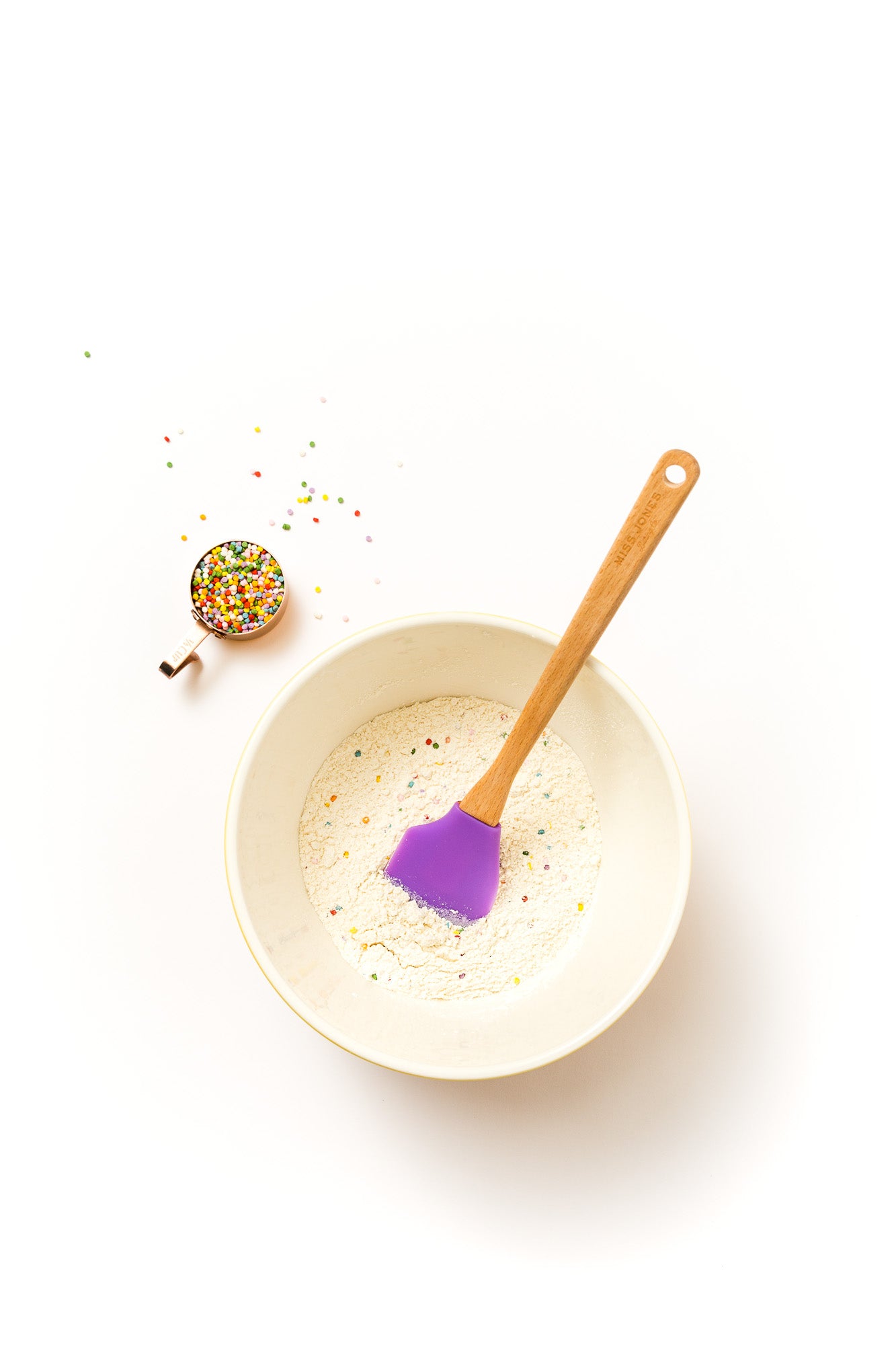 Image from above of Miss Jones Confetti Pop Cookie Mix in a mixing bowl with a purple spatula, next to a cup of rainbow sprinkles 