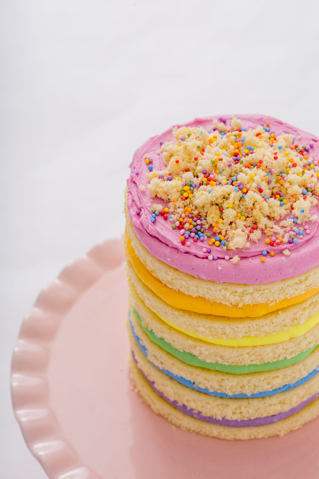 Image of the top of Miss Jones Baking Co Naturally Dyed Rainbow Frosted Layer Cake on a pink cake stand, covered in cake crumbles and rainbow sprinkles