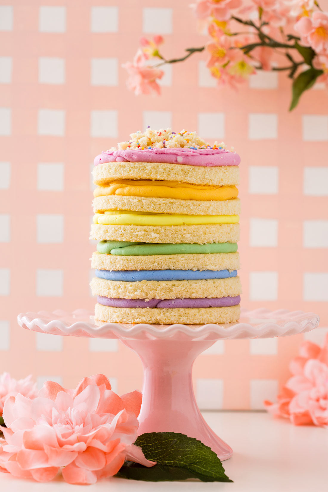 Image of the side of Miss Jones Baking Co Naturally Dyed Rainbow Frosted Layer Cake on a pink cake stand