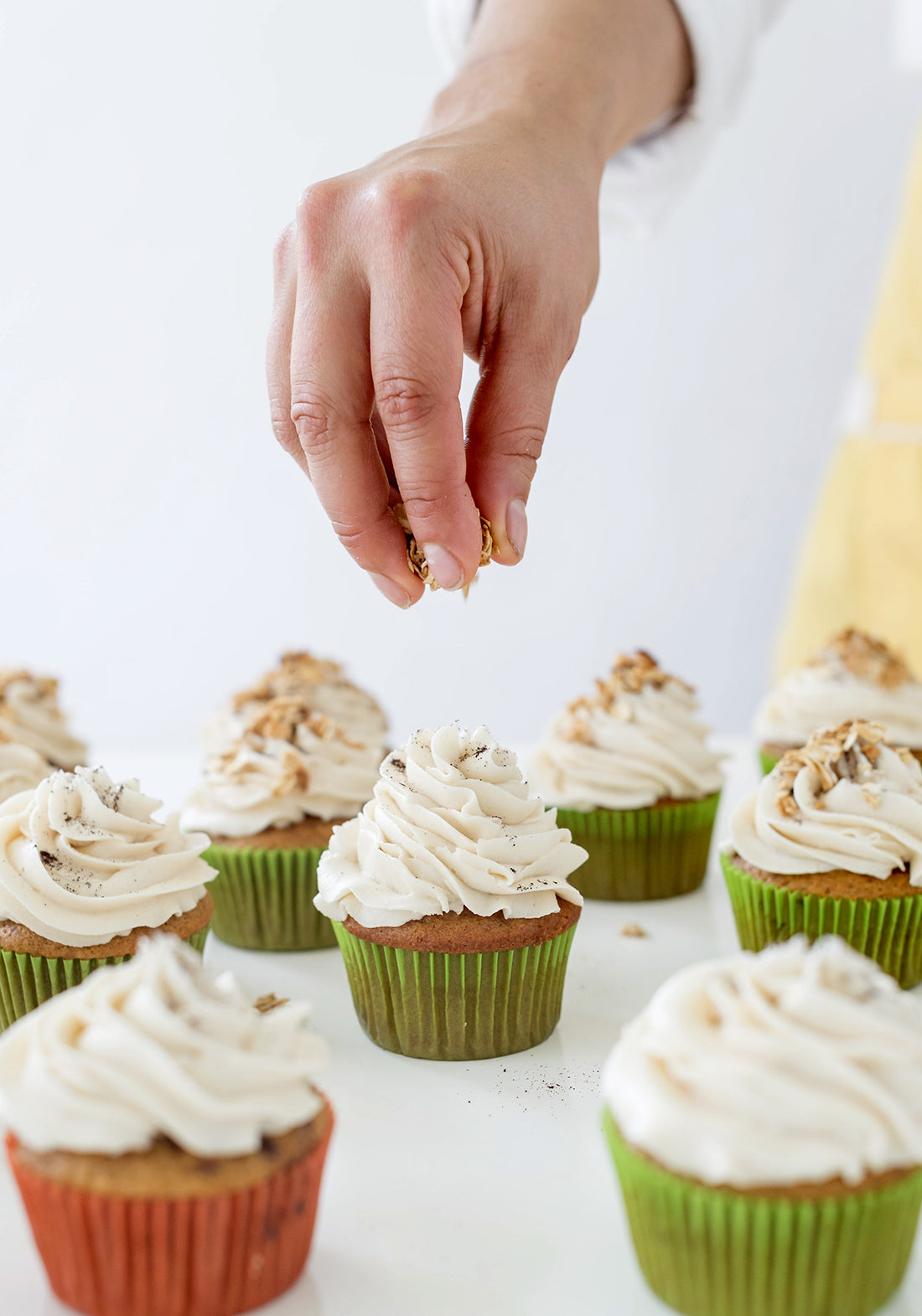 Image from the side of a hand sprinkling toppings on top of Miss Jones Baking Co Pumpkin Spice Latte (PSL) Cupcakes