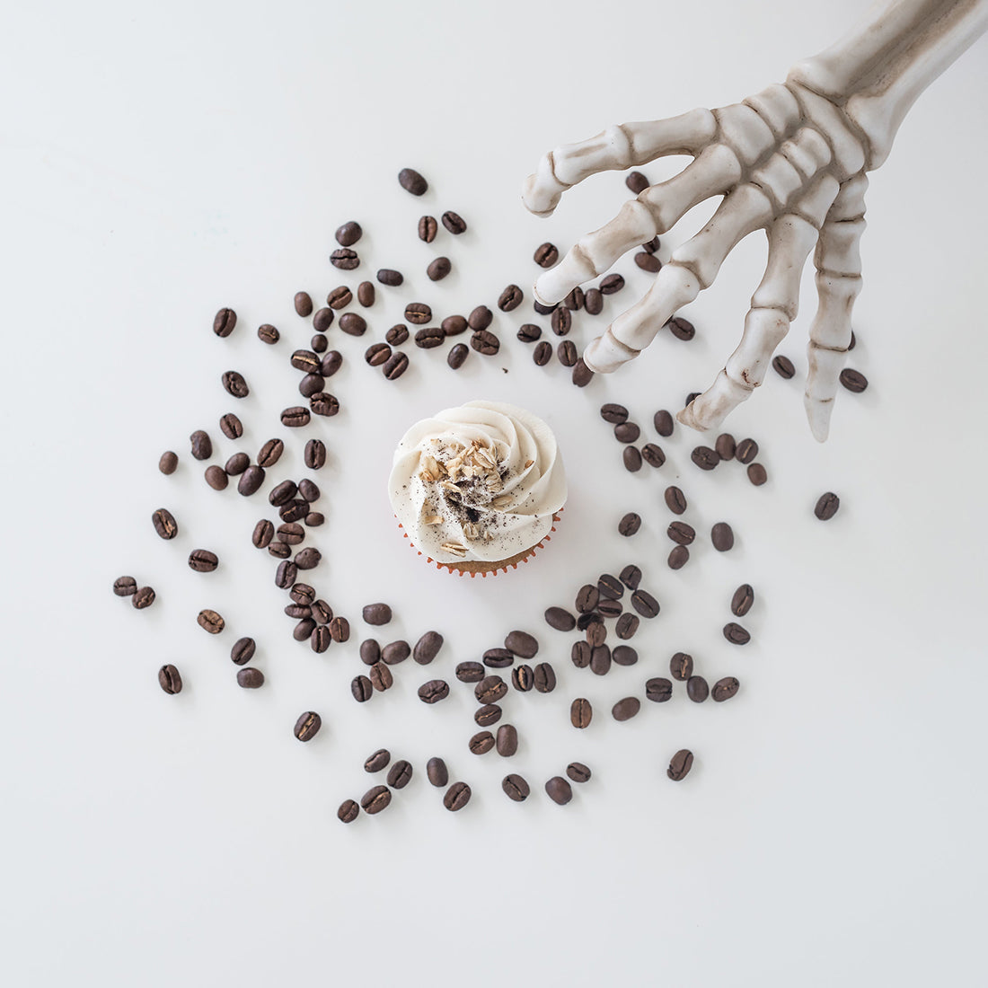 Image from above of a Miss Jones Baking Co Pumpkin Spice Latte (PSL) Cupcake surrounded by coffee beans next to a skeleton hand