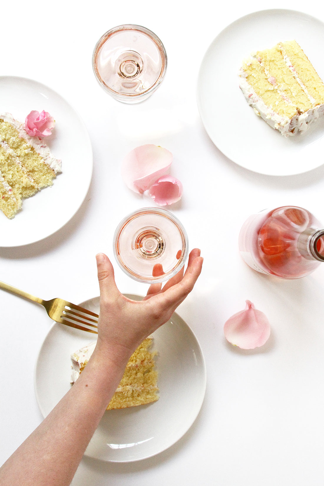 Image from above of three slices of Miss Jones Baking Co Rosé All Day Rose Cake next to a bottle of rosé, two glasses of rosé and rose petals