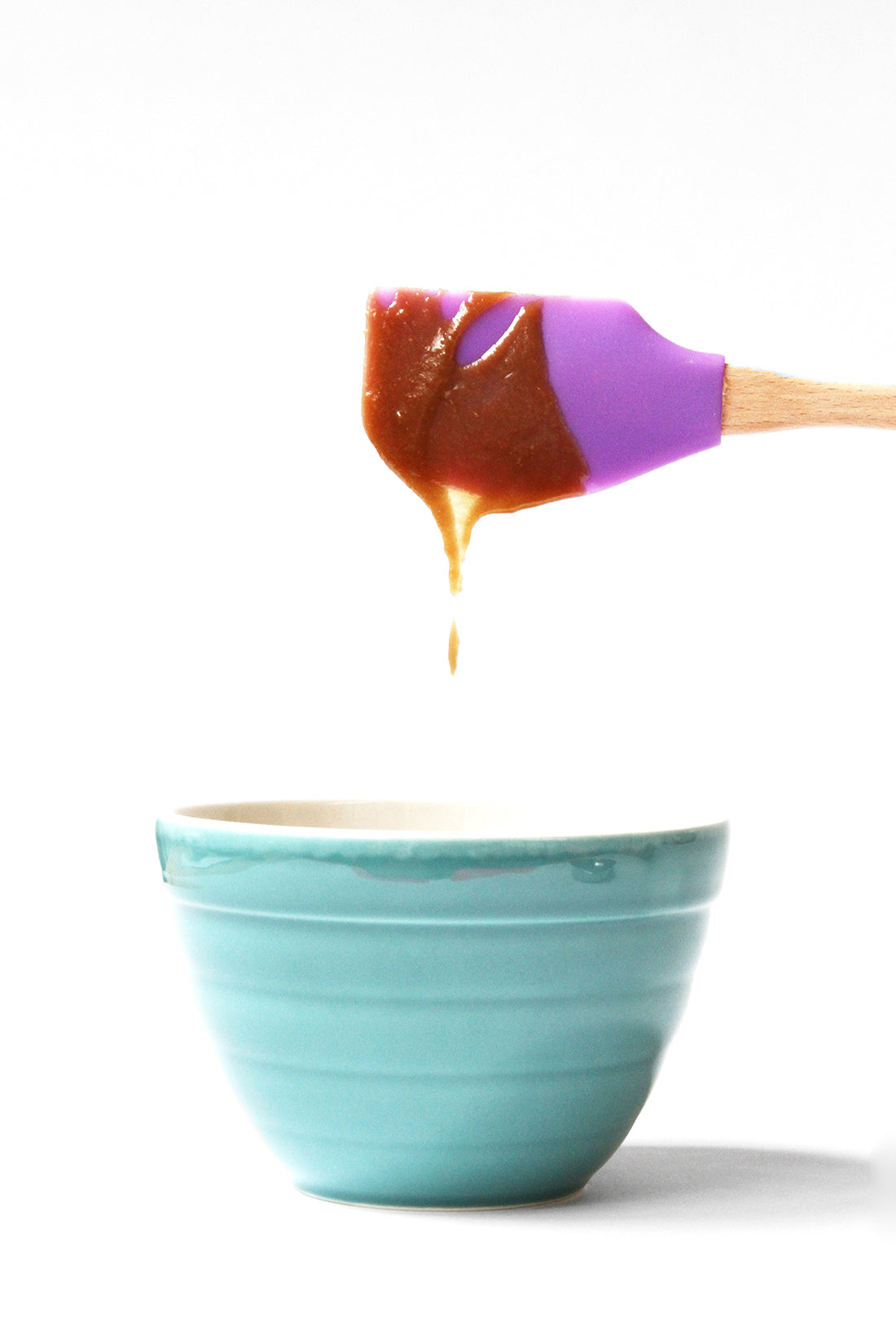 Image from the side of a purple spatula covered in toffee dripping into a blue mixing bowl for Miss Jones Baking Co Sticky Toffee Cakes