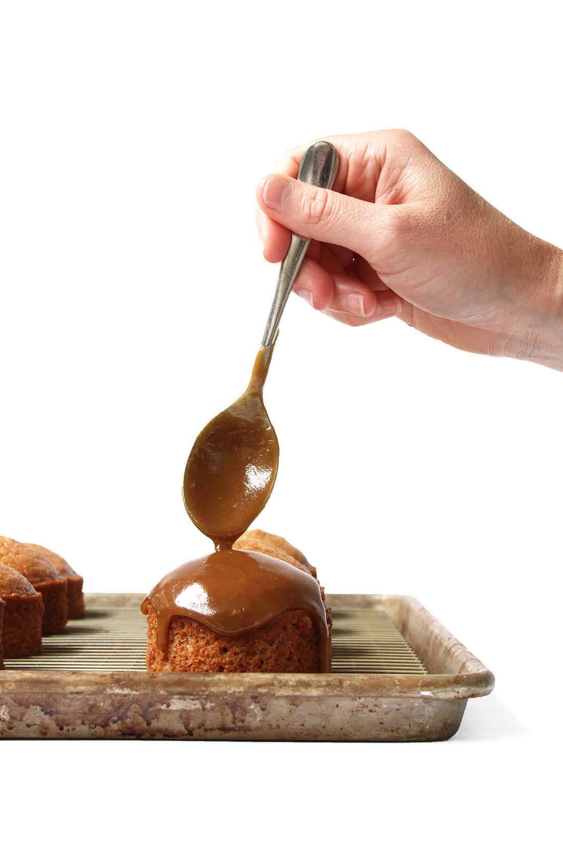 Image from the side of a hand holding a spoon drizzling toffee onto Miss Jones Baking Co Sticky Toffee Cakes on a baking sheet