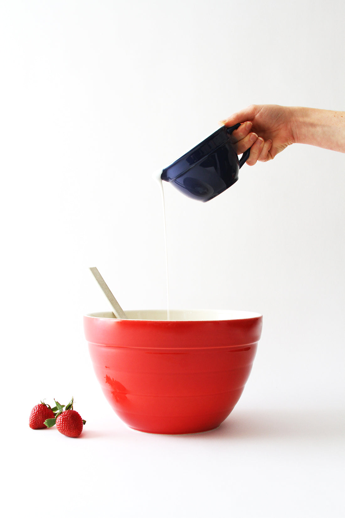 Image of cream poured into red mixing bowl next to two strawberries used for Miss Jones Baking Co California Strawberry Cake