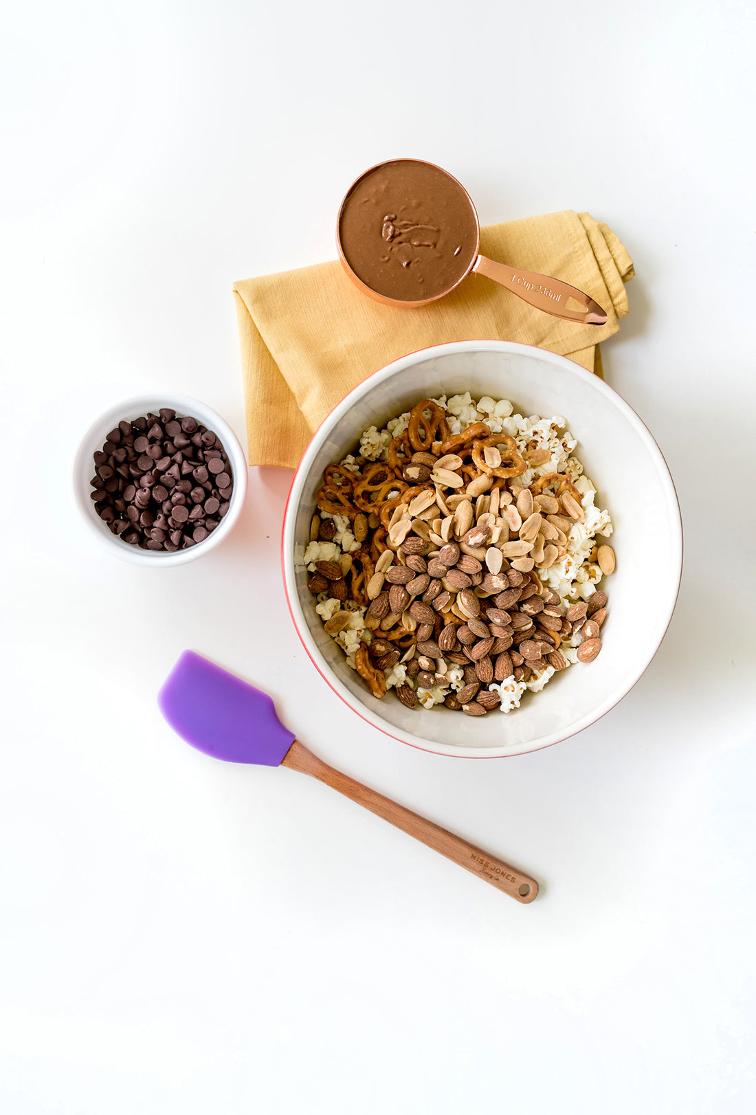 A bowl of snack ingredients mixed next to a purple spatula, a bowl of chocolate chips, and a cup of almond butter used for Miss Jones Baking Co Confetti Pop Puppy Chow Snack Mix Recipe