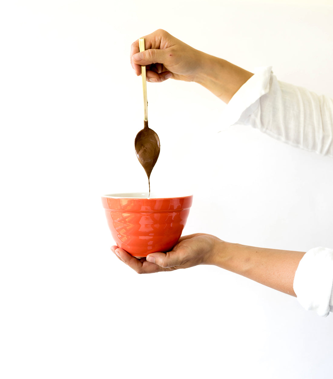 Image from the side of a hand holding a golden spoon dripping chocolate mixture into a red bowl for Miss Jones Baking Co Confetti Pop Puppy Chow Snack Mix