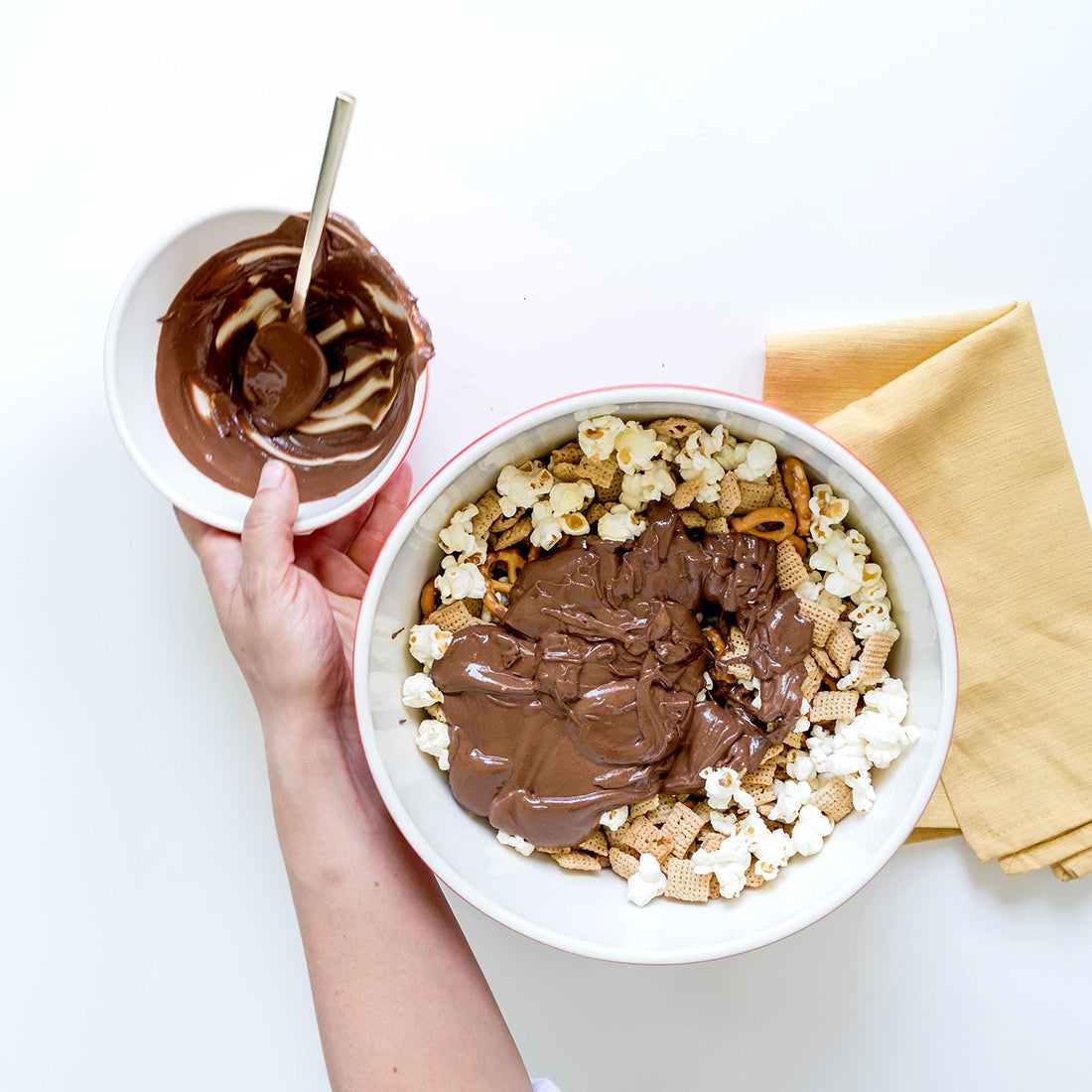 Image from above of a bowl filled with ingredients for Miss Jones Baking Co Confetti Pop Puppy Chow Snack Mix, topped with chocolate sauce next to another bowl of chocolate