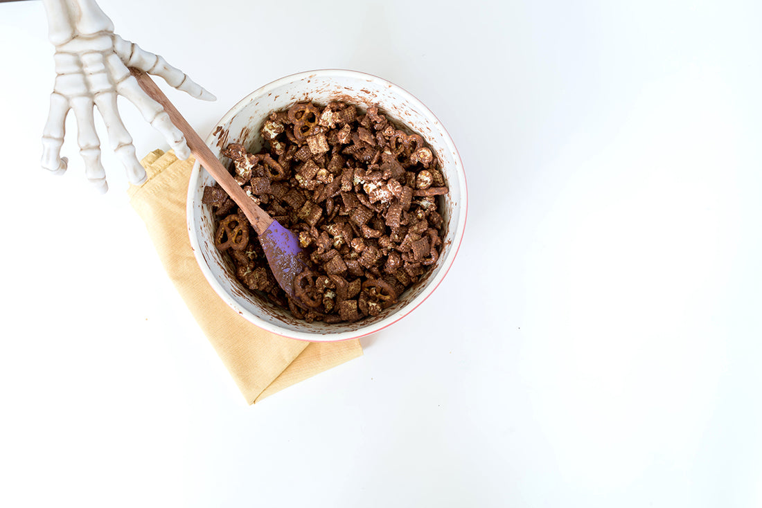 Image from above of a skeleton hand holding a purple spatula in a mixing bowl filled with chocolate covered ingredients for Miss Jones Baking Co Confetti Pop Puppy Chow Snack Mix
