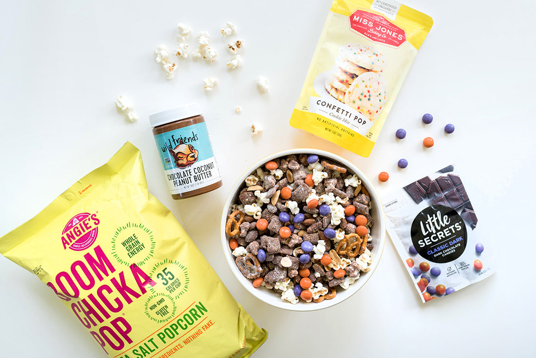 A bowl of Miss Jones Baking Co Confetti Pop Puppy Chow Snack Mix surrounded by a bag of Boom Chicka Pop Sea Salt Popcorn, a jar of Wild Friends Chocolate Coconut Peanut Butter, a bag of Miss Jones Confetti Pop Cookie Mix, and a bag of Little Secrets Classic Dark Dark Chocolate Candies