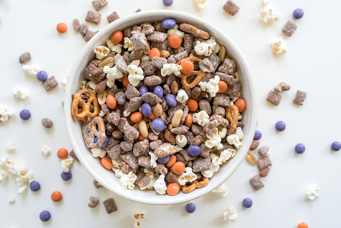 Close up image of a bowl of Miss Jones Baking Co Confetti Pop Puppy Chow Snack Mix surrounded by pieces of Miss Jones Baking Co Confetti Pop Puppy Chow Snack Mix