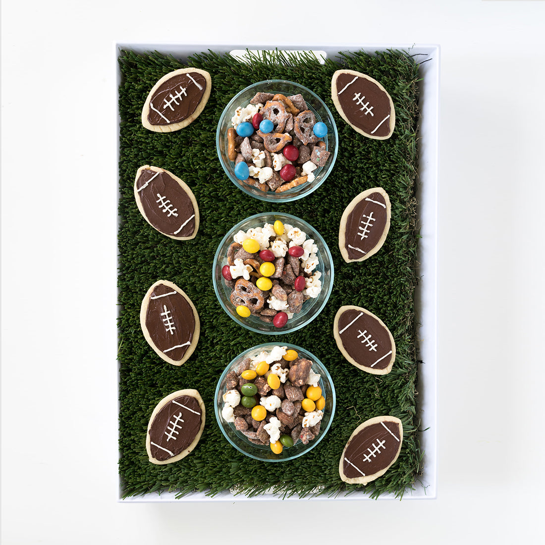 Image from above of three different bowls of Miss Jones Baking Co Confetti Pop Puppy Chow Snack Mix surrounded by football sugar cookies on a turf mat