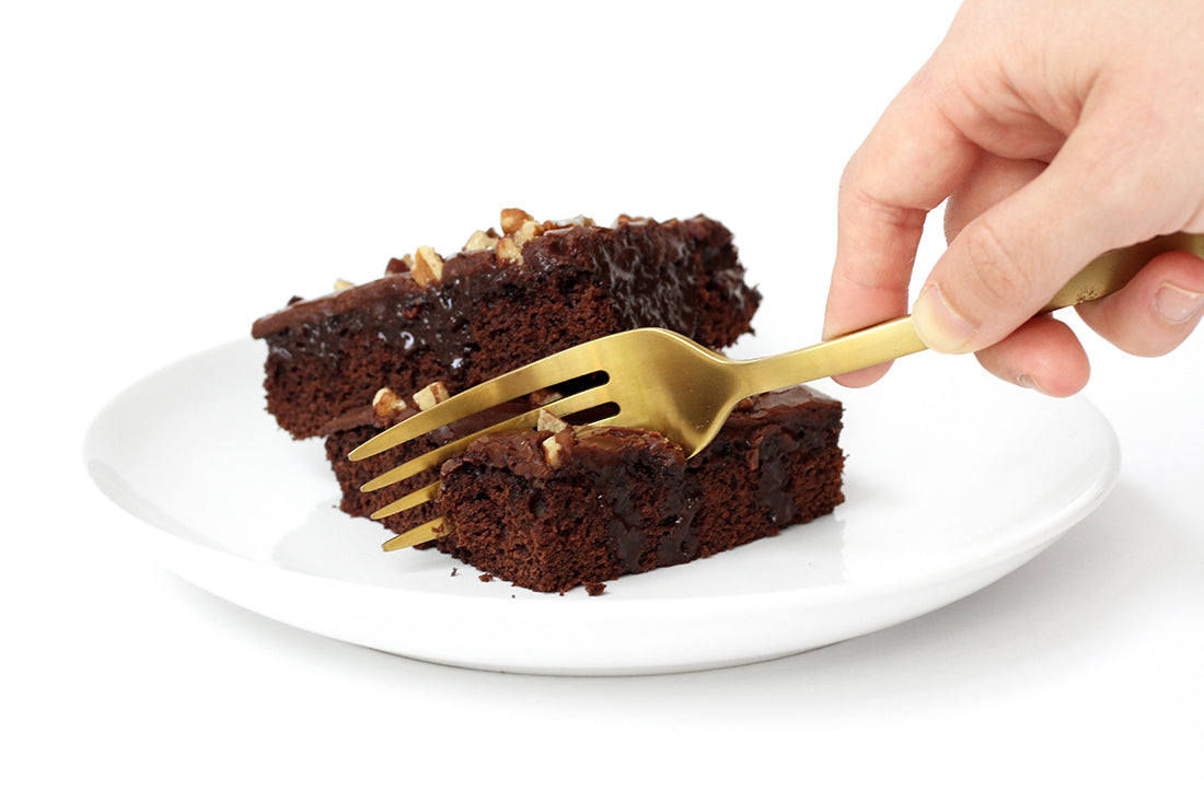 Close up image of two slices of Miss Jones Baking Co Sarah's Texas Sheet Cake on a white plate with a hand taking a bite of one with a gold fork