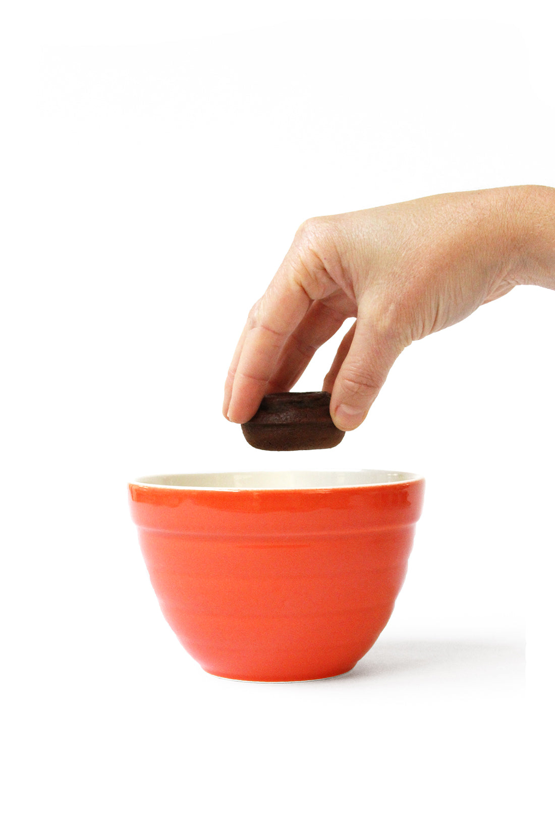 Image of the side of a hand dipping an unfrosted Miss Jones Baking Co Thin Mint Donuts into a red mixing bowl