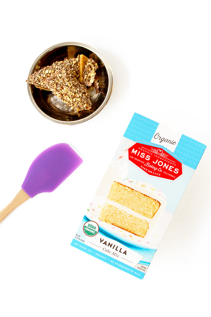 Image from above of a box of Miss Jones Vanilla Cake Mix, a bowl of toffee, and a purple spatula used for Miss Jones Baking Co Sarah's Toffeedoodles recipe