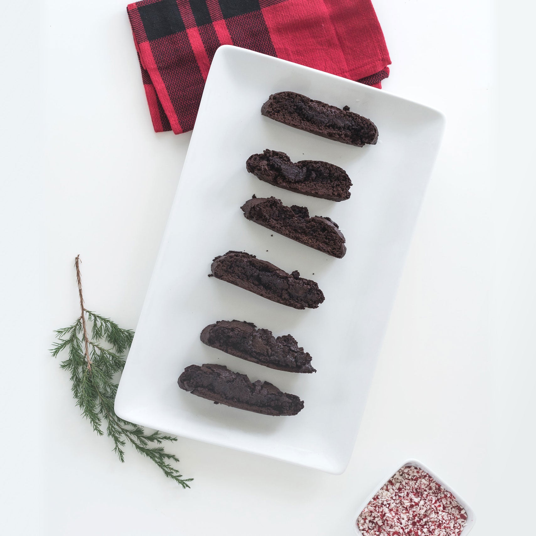 Image of six Miss Jones Baking Co Easy Chocolate Peppermint Cake Mix Biscotti cookies on a white platter next to a red towel and a bowl of crushed peppermint candies