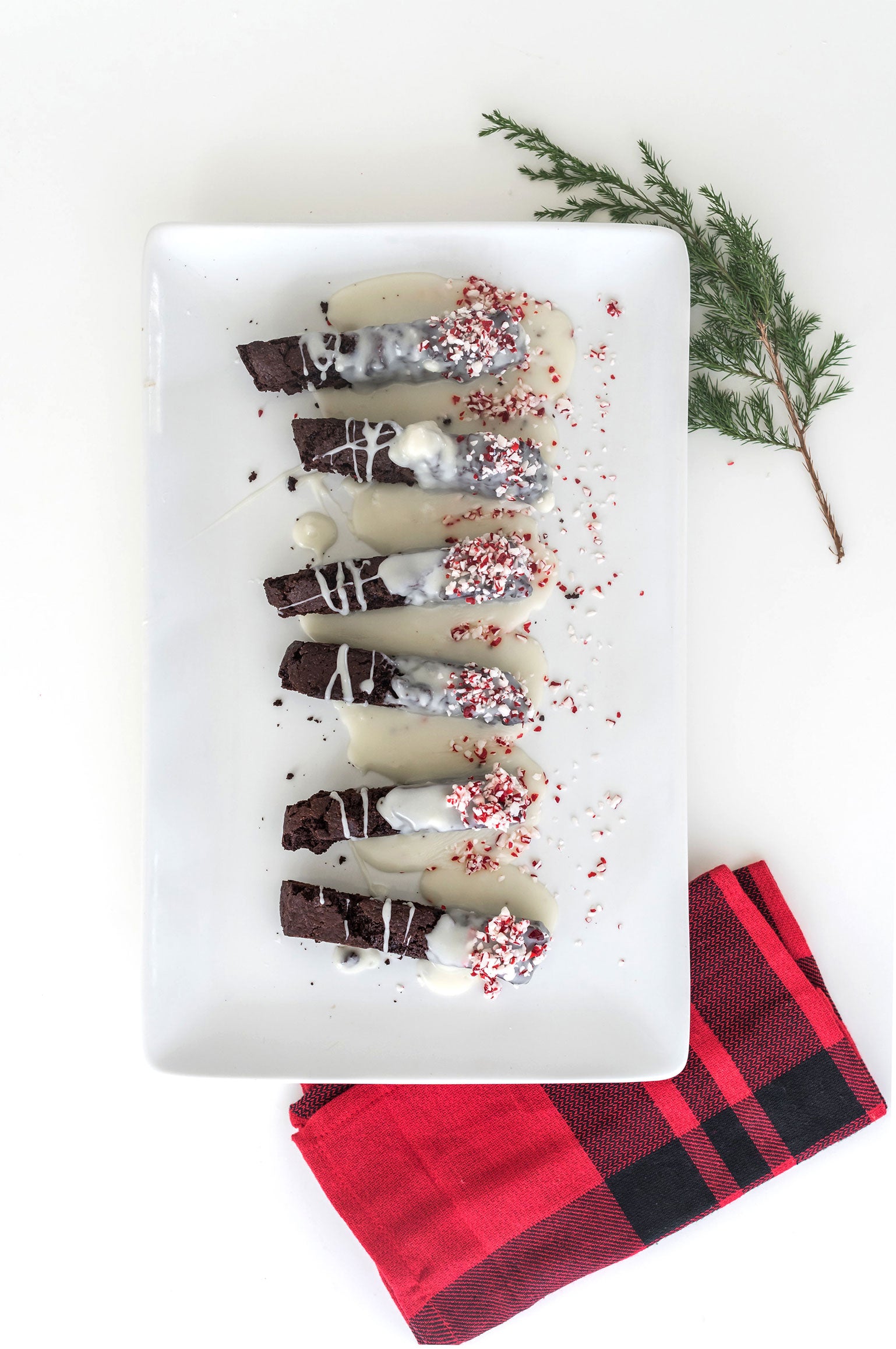 Image from above of six Miss Jones Baking Co Easy Chocolate Peppermint Cake Mix Biscotti cookies on a white plate next to a red towel and a sprig of Christmas tree