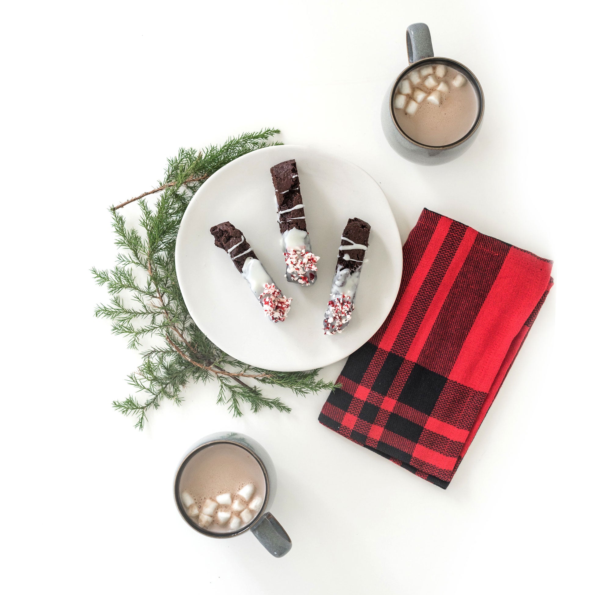 Image from above of three Miss Jones Baking Co Easy Chocolate Peppermint Cake Mix Biscotti cookies on a white plate next to two mugs of hot chocolate with mini marshmallows, a red towel, and a sprig of Christmas tree