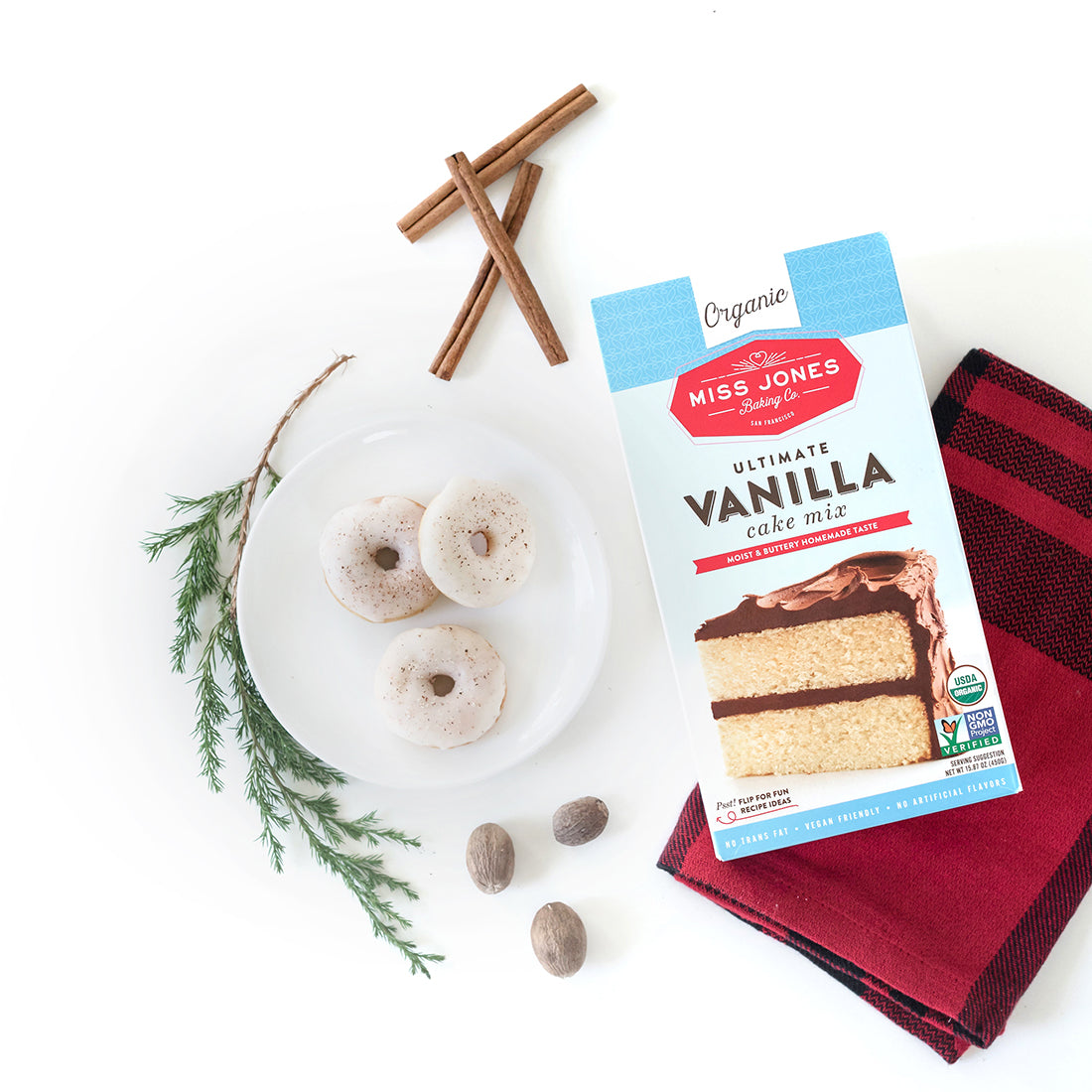 Image of three Miss Jones Baking Co Easy Holiday Eggnog Mini Donuts on a white plate next to a box of Miss Jones Vanilla Cake Mix, three nutmegs, three cinnamon sticks and a sprig of a conifer branch