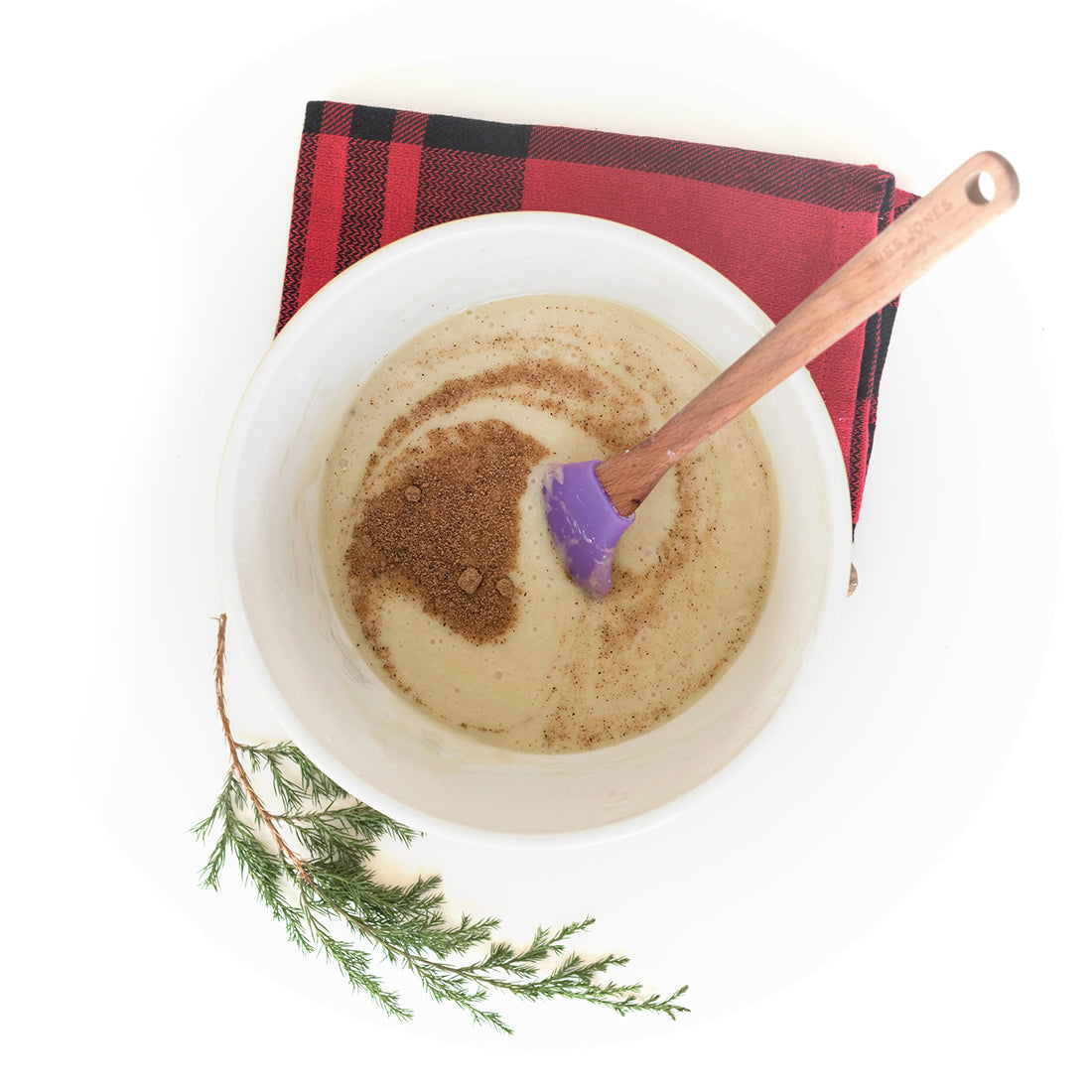 Image from above of Miss Jones Baking Co Easy Holiday Eggnog Mini Donuts batter in a mixing bowl with a purple spatula next to a red towel and a sprig of a conifer branch