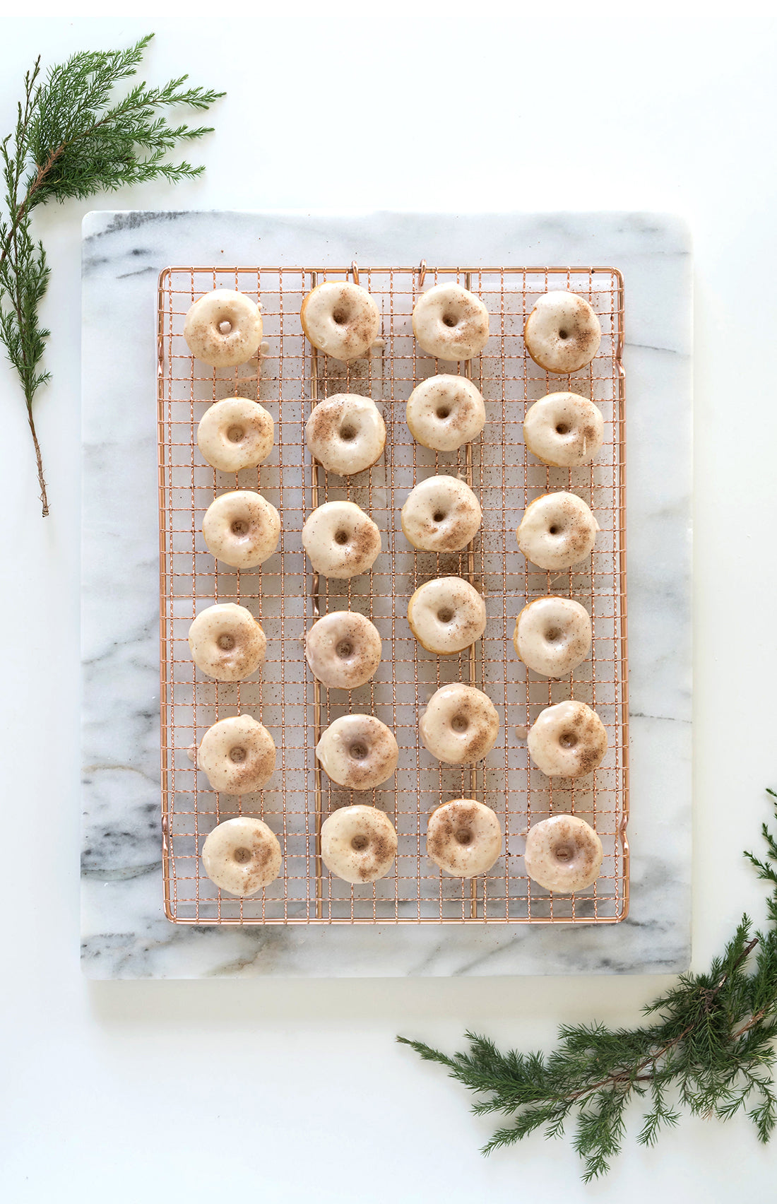 Image of the top of 24 Miss Jones Baking Co Easy Holiday Eggnog Mini Donuts on a copper. baking rack next to two sprigs of conifer branches