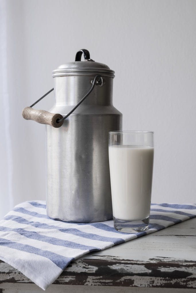 A metal jug of milk on a table, next to a glass of milk.