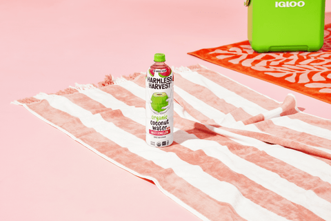 a bottle of harmless harvest watermelon organic coconut water on a pink and white striped beach towel next to a green cooler