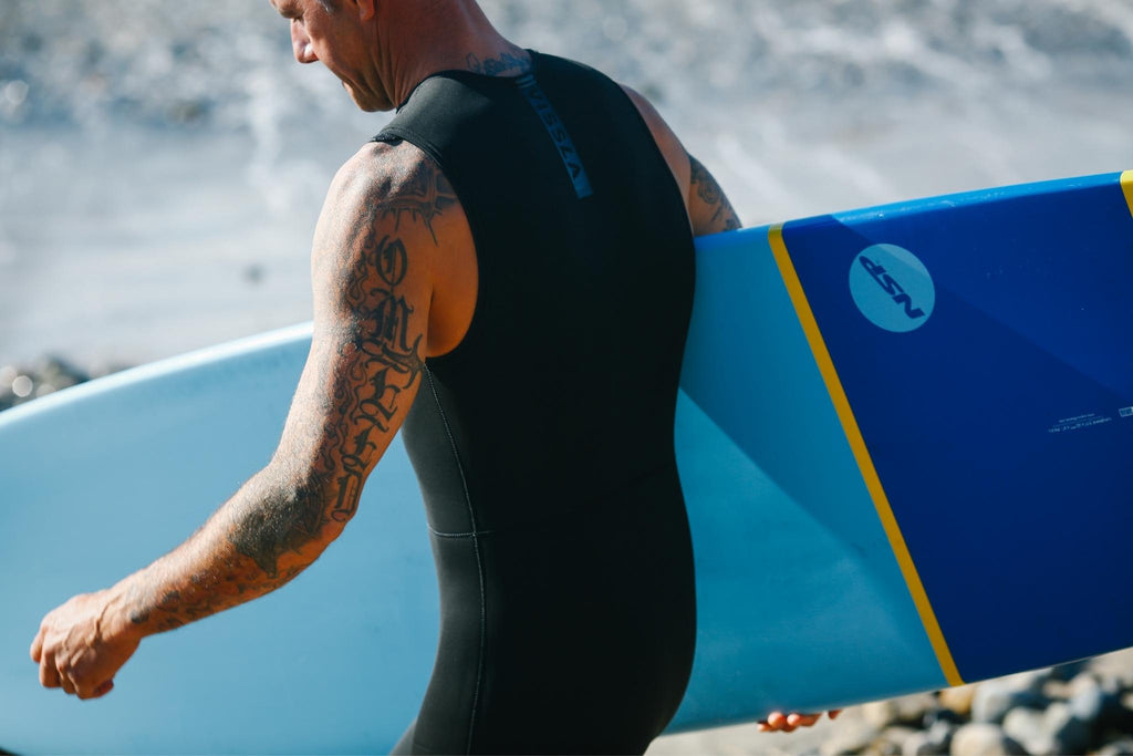 Man in wetsuits waling on shore carrying his NSP Surfboard