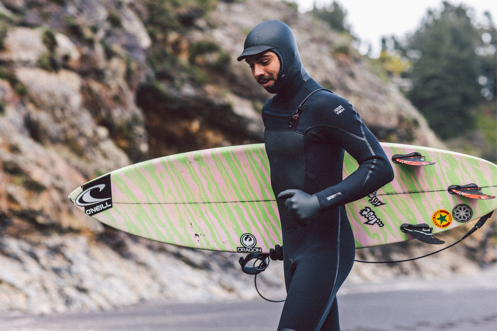 Man in wetsuits with gloves and hood walking on the beach carrying his surfboard