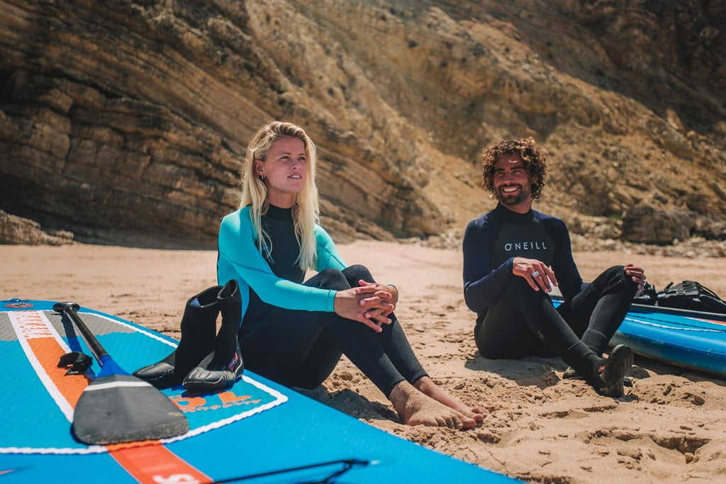 Couple sitting on the beach beside Stand up paddle boards while wearing O'Neill Reactor wetsuits