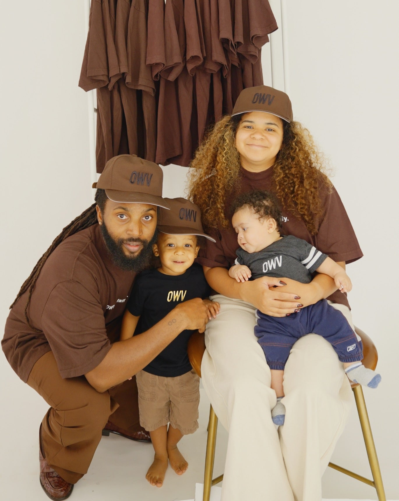 O'Shea, on the left, with his family. O'Shea is wearing an all brown outfit, including a hat with the initials OWV embroidered on it. He's holding his older son, who is wearing an OWV black shirt and a brown hat. On a chair next to them is his wife, whose brown/blonde curly hair is springing out from underneath another brown OWV hat. She's smiling, and holding their infant son, who's wearing a blue outfit and looking at the toddle on the floor next to him. Behind them, a rack of brown shirts hang neatly.