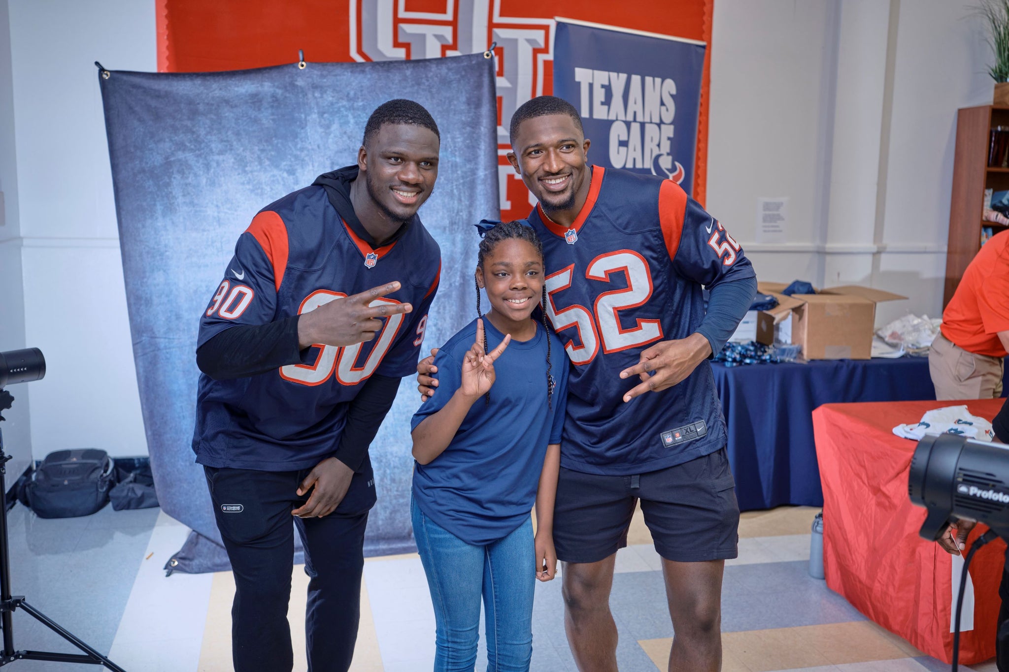 A girl with braids and throwing up a peace sign poses for a photo between two Texans players in their jerseys.