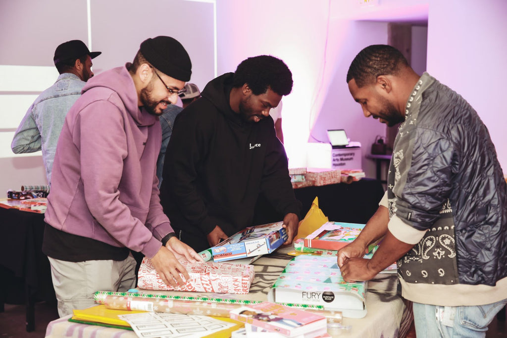 Three men stand at a table wrapping gifts.