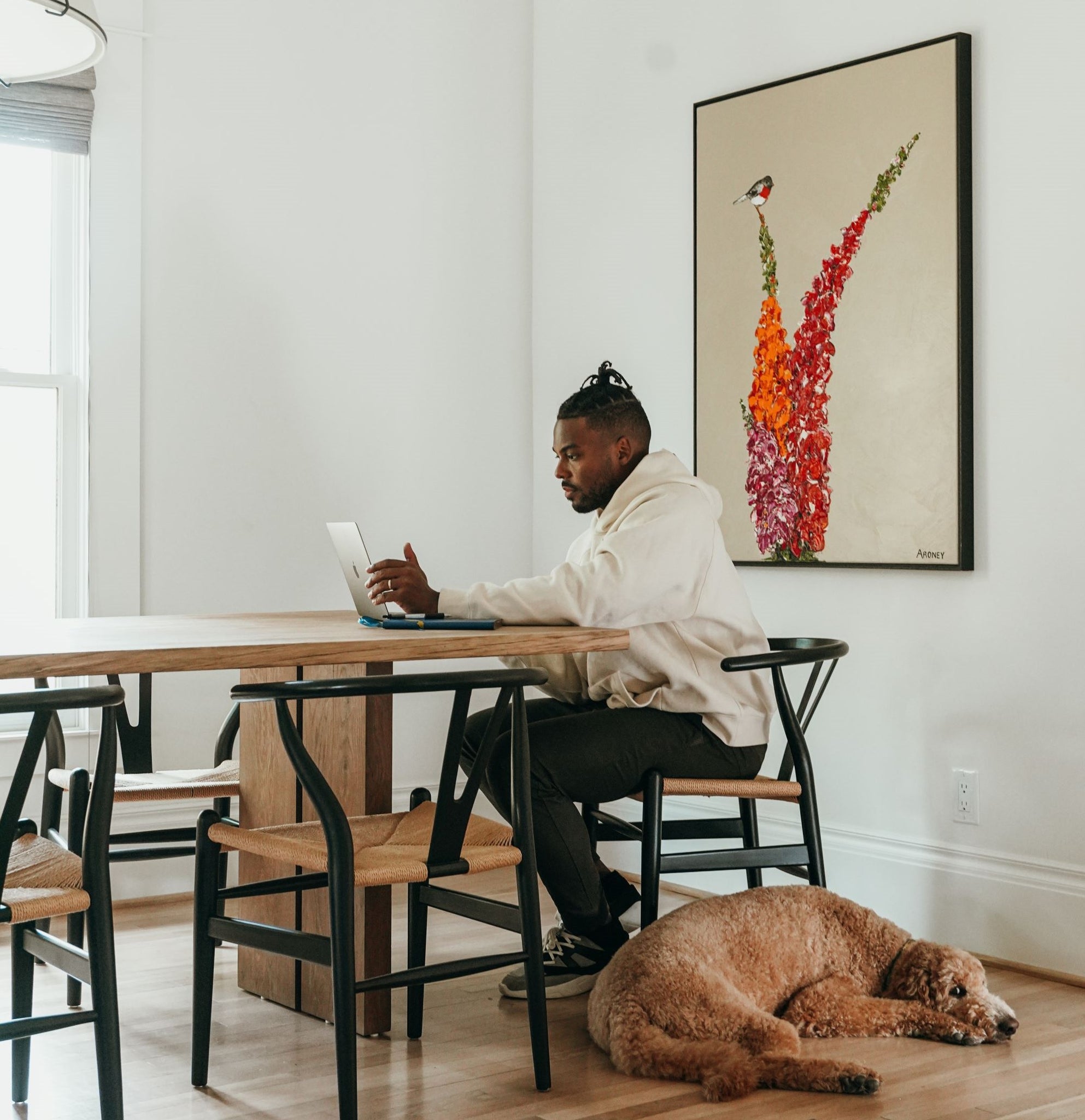 Troy sits at his dining room table looking at a laptop. His dog, a fluffy brown labradoodle, is curled up beneath him on the ground. A large painting of a bright red bird is behind him on the wall.