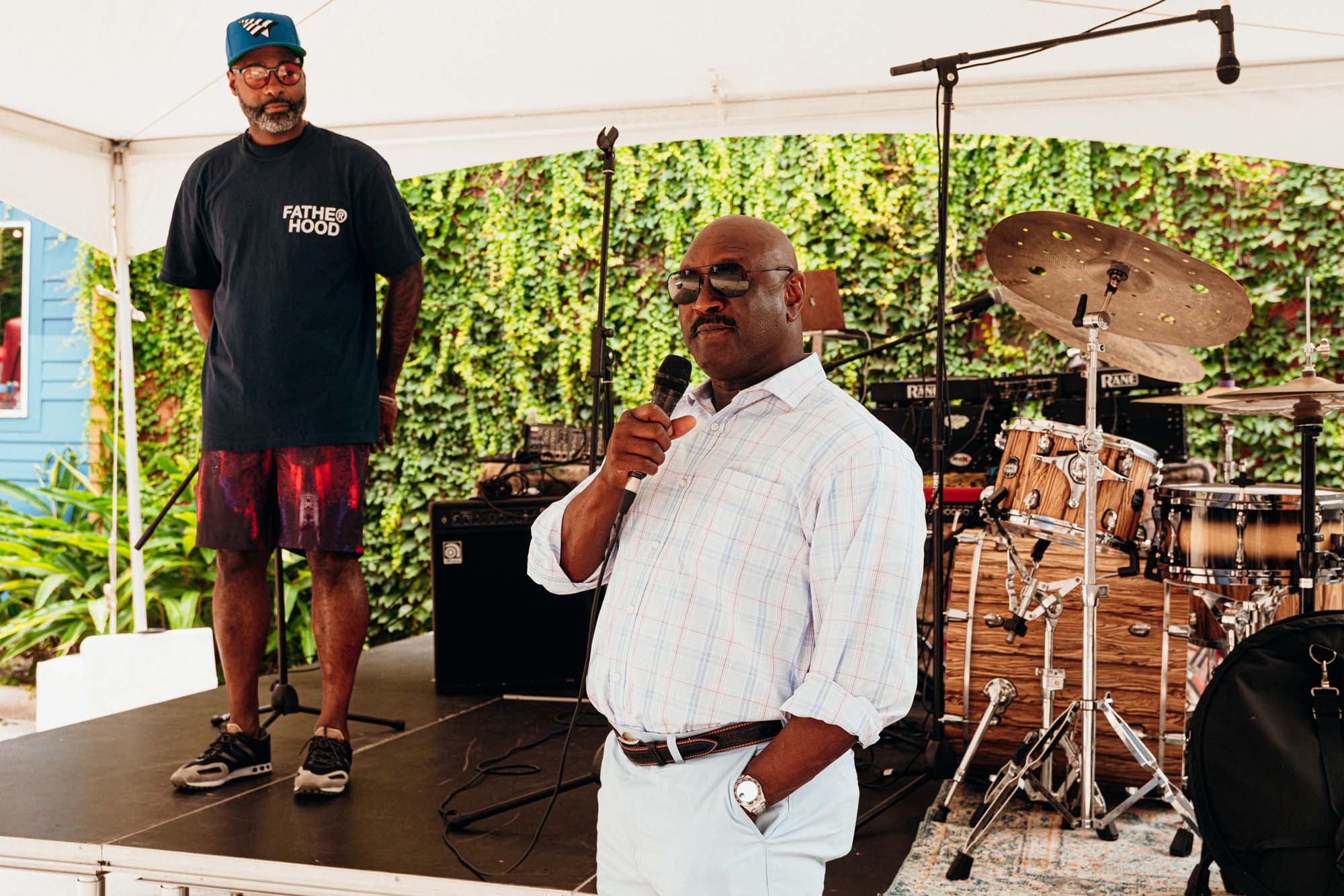 James Harris, a Black man wearing a white button up collared shirt and light blue dress pants, speaks into a microphone. He has one hand in his pocket. Kevin, in a black T-shirt and a ball cap, stands behind him on a stage. It's a sunny day. On the stage also are drums and a microphone stand.